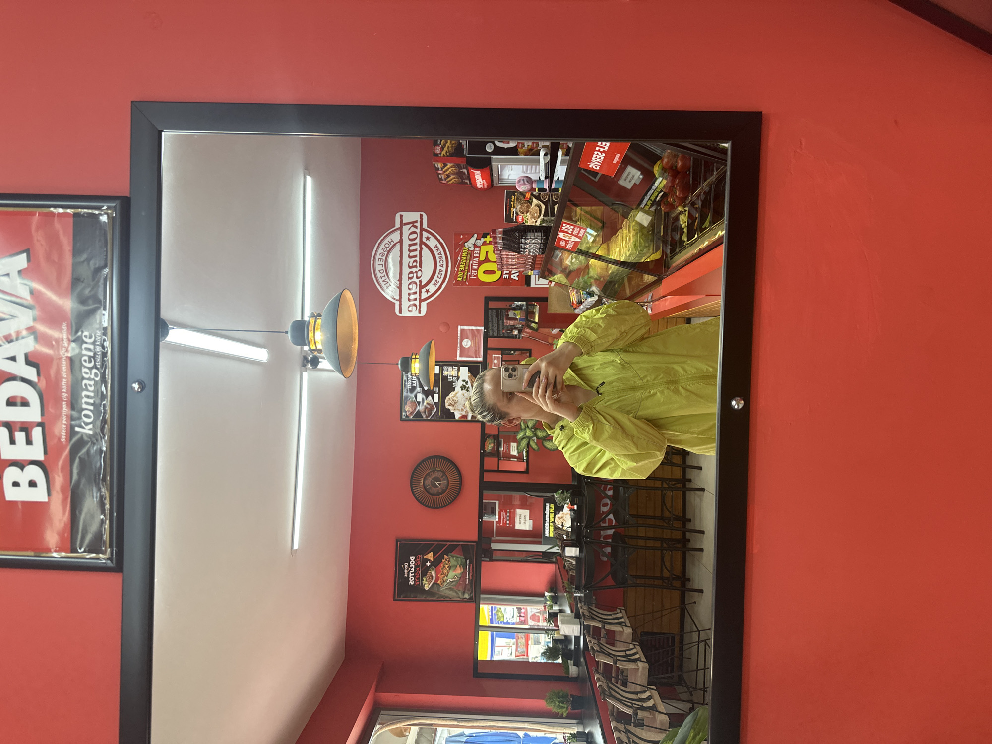 Here is a concise description of the scene:
The image shows a reflection in a mirror of a person taking a selfie in what appears to be a restaurant. The restaurant has red walls and features the "Komagene" brand, known for çiğ köfte (raw meatballs). Various posters and advertisements are displayed on the walls. The reflection shows tables and chairs in the restaurant. A sign indicating "Bedava komagene" is also seen. The location is likely Fethiye, Turkey, given the common presence of Komagene restaurants in the area.
