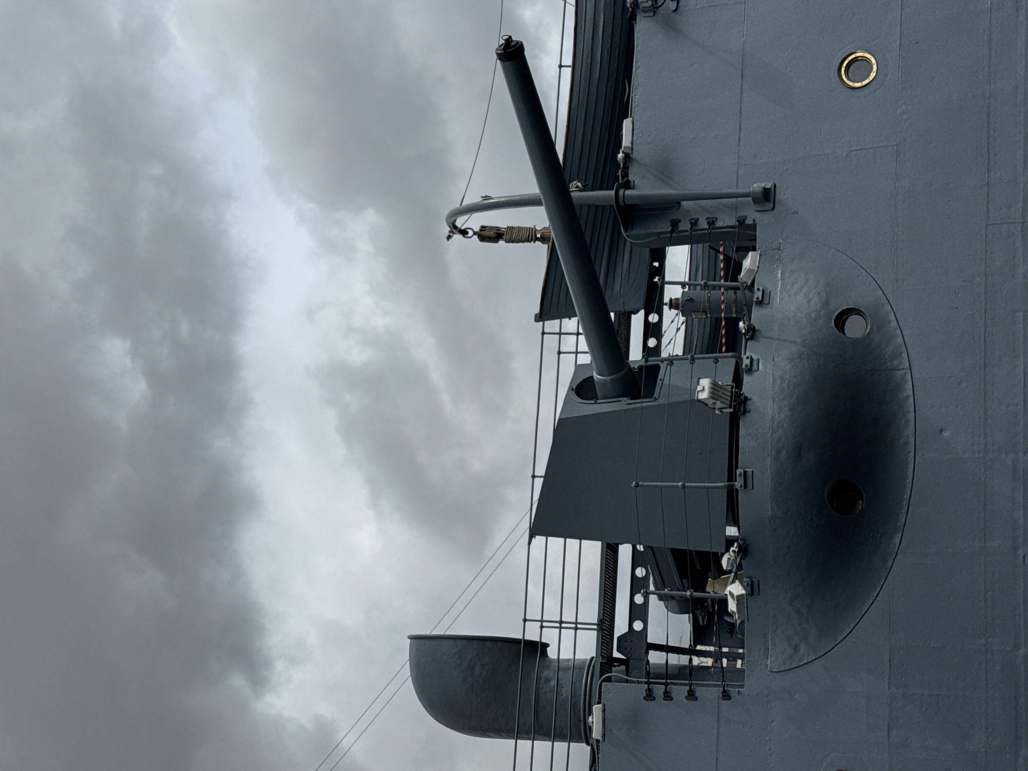 The image shows a portion of a naval vessel, likely a warship or a museum ship. The dominant features are a large gun turret with a prominent gun barrel, and part of the ship's hull. The ship is painted in shades of gray. The background consists of a cloudy, overcast sky. The location is potentially Saint Petersburg, Russia, if the vessel is a museum ship docked in the city.