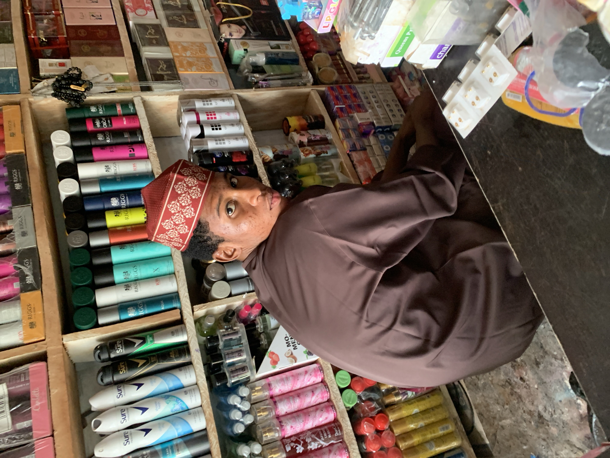 The image shows a young man wearing a brown traditional outfit and a red and white embroidered cap, facing away from the camera, in what appears to be a small shop in Abuja, Nigeria. He is sitting down and looking over his shoulder towards the camera. The shop is filled with shelves stacked with various products, including deodorants, perfumes, lotions, and other cosmetic items. The shelves are made of wood and the products are arranged in an organized manner. The lighting in the shop is adequate, and the overall scene suggests a typical retail environment in a Nigerian city.