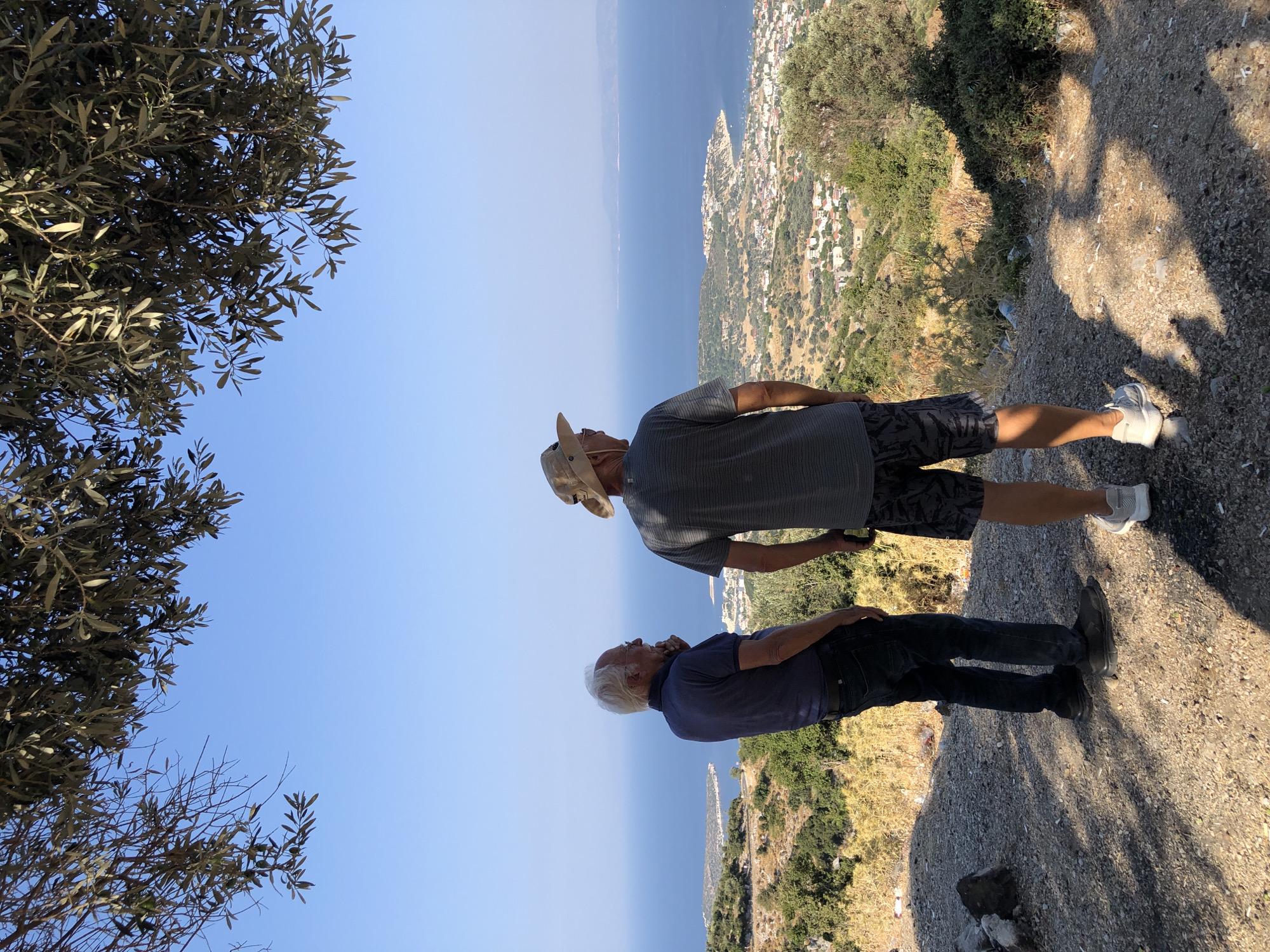 Two individuals are captured from behind, standing on a rocky, elevated terrain overlooking a coastal town and the Aegean Sea. The person on the left, an older man with white hair and glasses, wears a dark blue polo shirt and dark jeans. He stands with his right hand on his chin, gazing out at the vista. To his right, a younger man in a grey t-shirt, patterned shorts, and a tan hat faces the same direction. He wears white athletic shoes, while the older man sports dark slip-on shoes. Their focus is directed towards a peninsula with a densely populated town nestled at its base, leading into the expansive, calm blue sea. The background is characterized by a clear, bright blue sky, with the upper portion of the frame occupied by the branches and leaves of an olive tree. The terrain they stand on is dry and sparse, dotted with rocks and dry vegetation, suggestive of a Mediterranean climate. The overall impression is one of quiet observation and appreciation of a scenic landscape. The town's architecture appears to be a mix of red-tiled roofs and white-washed buildings, typical of coastal Aegean settlements. The presence of a tree with olive leaves indicates the location is likely within the Mediterranean region, consistent with Karaburun, Turkey, known for its olive groves and coastal beauty.