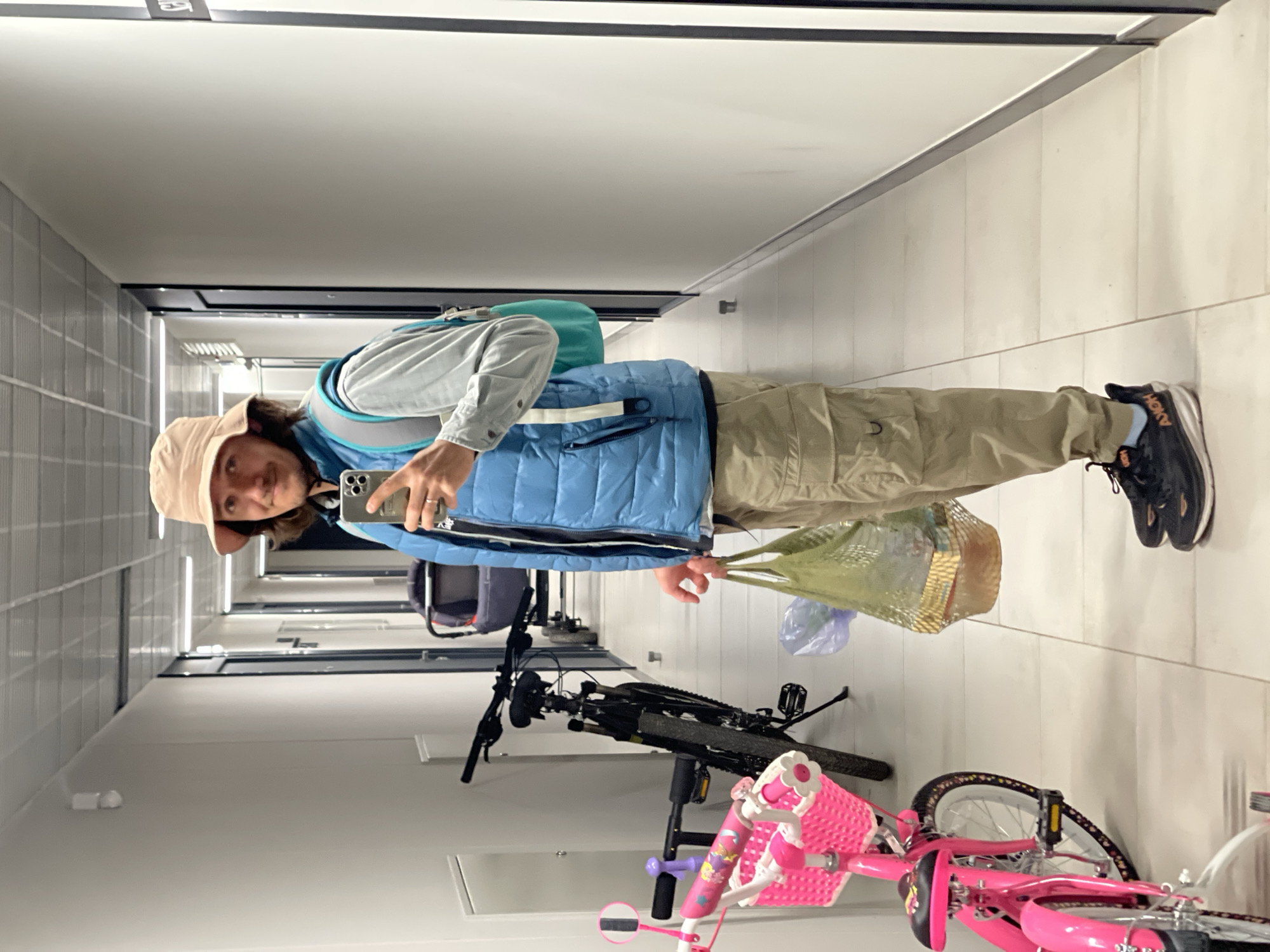 A person wearing a tan bucket hat, blue vest, light grey button-down shirt, tan pants, and black Hoka sneakers is standing in a hallway with tiled walls. They are holding a smartphone in their left hand, seemingly taking a selfie, and a mesh shopping bag in their right hand. The bag appears to contain some groceries and a plastic bag. A turquoise backpack is visible behind the person.
In the background, two bicycles are parked against the wall. One is a black mountain bike, and the other is a pink children's bicycle with a basket. The hallway has white tiled floors and white walls with black trim along the ceiling. The hallway extends into the distance with visible doorways on the left-hand side.