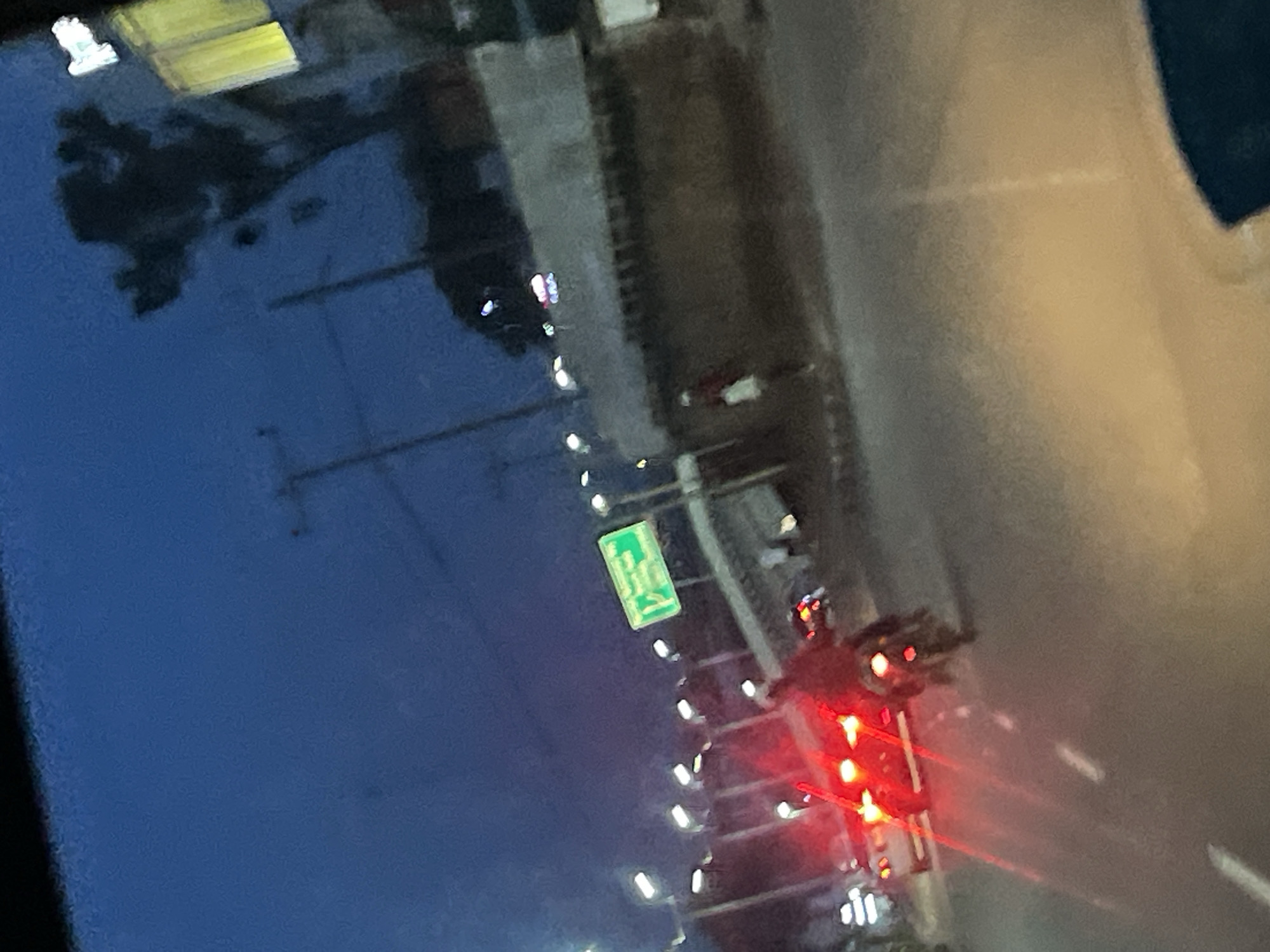 The image depicts a nighttime scene on a street in Jos, Nigeria. A red car with its brake lights illuminated is in the foreground, partially obscured by motion blur. To the right of the car, a motorcycle rider is also visible, with the motorcycle's headlight emitting a faint glow. The street appears to be paved and shows lane markings. In the background, a green directional sign with white text is mounted on a pole. The sky is a deep blue, indicating twilight or early night. Buildings, streetlights, and a silhouette of a tree are also present. The overall impression is one of moderate urban activity during the evening.