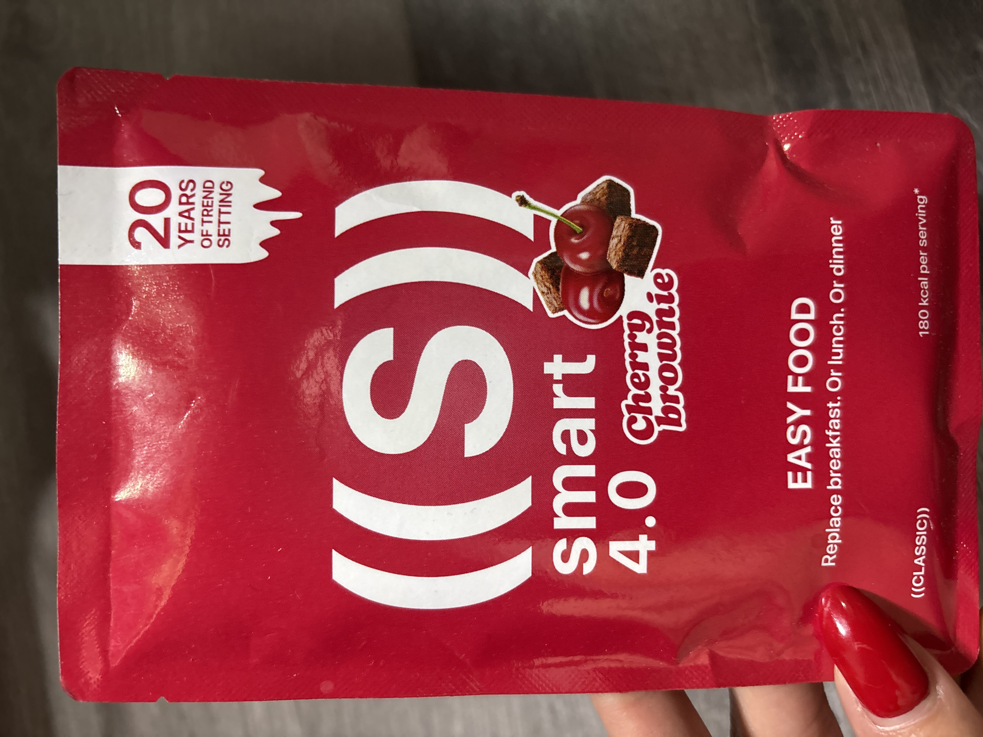 The image displays a close-up, eye-level shot of a red pouch with white text and graphics. The pouch is likely a food product. Dominating the upper half of the pouch is a large, white graphic that appears to be two stylized parentheses enclosing the letter "S". Below this, the word "smart" is printed in white, followed by "4.0" in white. To the right of "4.0", the words "Cherry brownie" are printed in a stylized white font.
In the lower right quadrant of the pouch, there is a graphic featuring two cherries and three small brownie pieces. To the top right of the pouch, a white tab-like design contains the text "20 YEARS OF TREND SETTING" in black and red lettering.
The bottom section of the pouch has the words "EASY FOOD" in large white capital letters, followed by smaller white text that reads "Replace breakfast. Or lunch. Or dinner". At the very bottom, there are two lines of text: "((CLASSIC))" on the left and "180 kcal per serving*" on the right, both in white.
A person's hand with bright red nail polish is visible on the left side of the image, holding the pouch. The background is a blurry, textured surface that appears to be a table or countertop. The lighting is even, with some reflections on the glossy surface of the pouch. The product appears to be new and undamaged, aside from some minor creases in the pouch material.