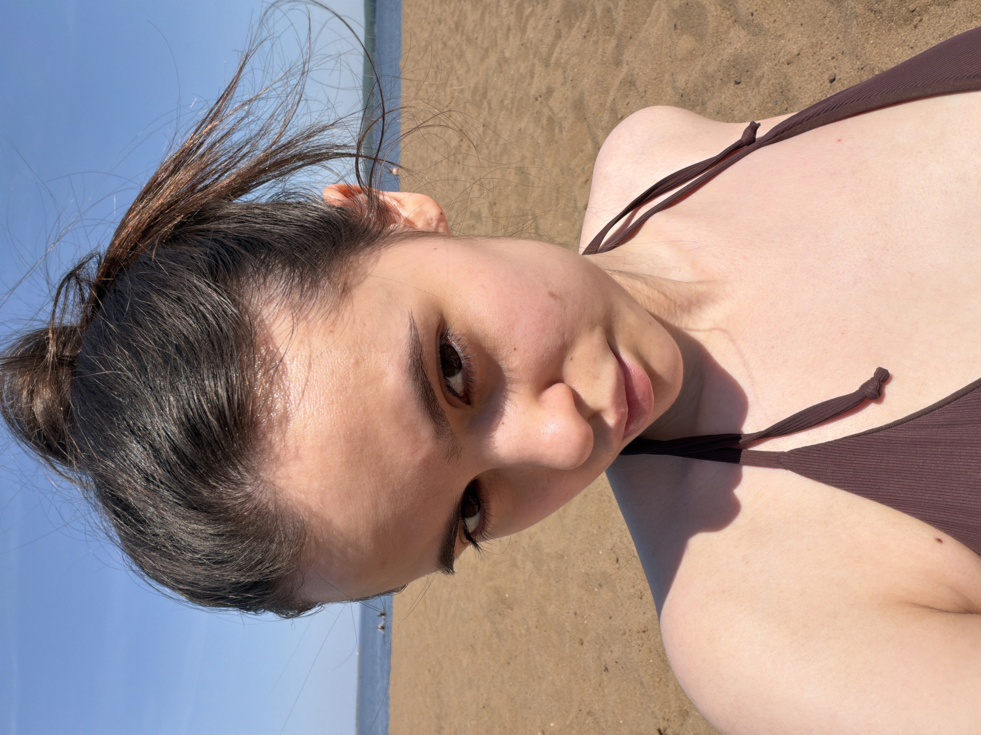 A woman with dark hair pulled into a bun is lying on a sandy beach. She is wearing a brown bikini top. The background shows the sea and sky, suggesting a beach setting. The photo was taken outdoors in daylight. Given the prompt's specification, this could be Chaikovsky, Russia.
