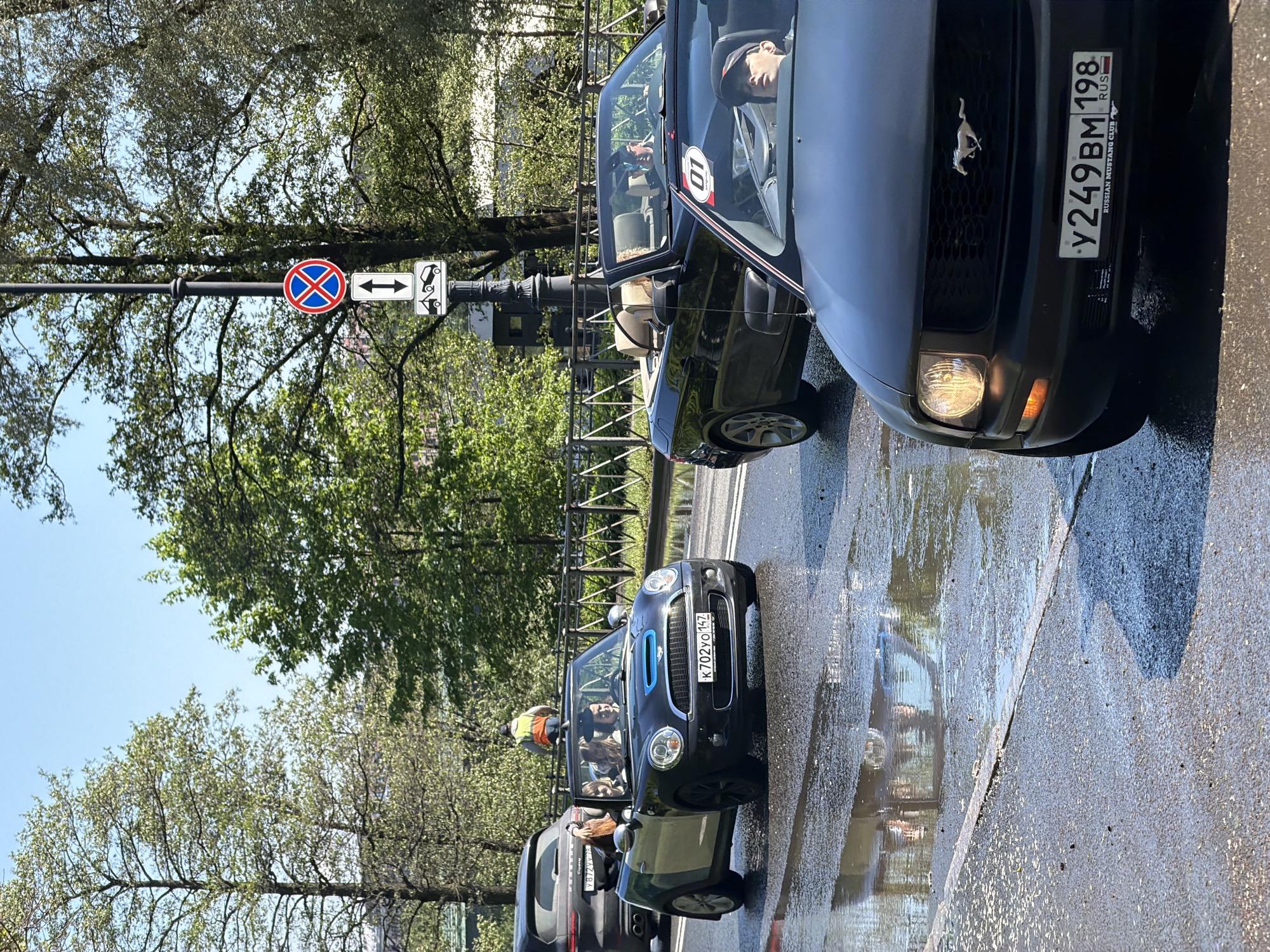 The image captures a street scene in Saint Petersburg, Russia. The foreground is dominated by a wet, cracked asphalt road reflecting sunlight and vehicles. On the right side of the frame is a black Ford Mustang with a visible license plate: Y249BM 198 RUS, indicating it's registered in Russia and possibly part of the Russian Mustang Club. The Mustang's front right headlight is on.
Behind the Mustang is a black convertible with the number "10" on its door, and the driver is visible inside. Adjacent to it is a black Mini Cooper with the license plate K702YO147, occupied by several people. A dark SUV is seen behind the Mini Cooper.
In the background, there are trees with green foliage and a blue sky visible through the branches. A street sign is mounted on a pole, indicating "no parking" and directing traffic in both directions. A metal fence separates the road from the trees. The lighting suggests it's a bright, sunny day.