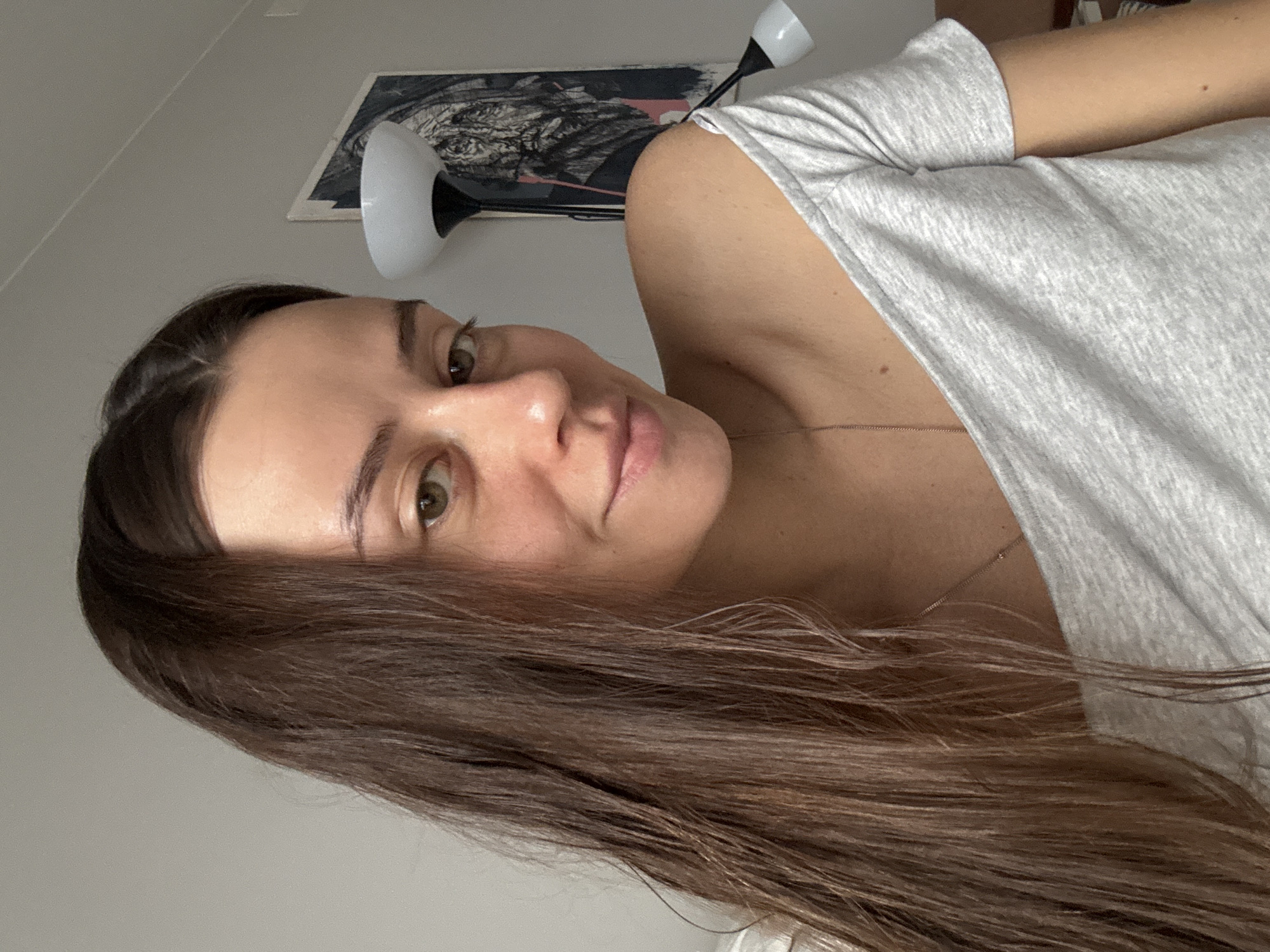 A woman with long brown hair and light olive skin is the subject of this indoor selfie. She's wearing a gray T-shirt that is slightly off-the-shoulder. Her eyes are brown, and she's looking at the camera with a slight smile. In the background, there's a wall lamp and a piece of abstract art. There is no discernible information from the image that indicates the location is Krasnoyarsk, Russia.