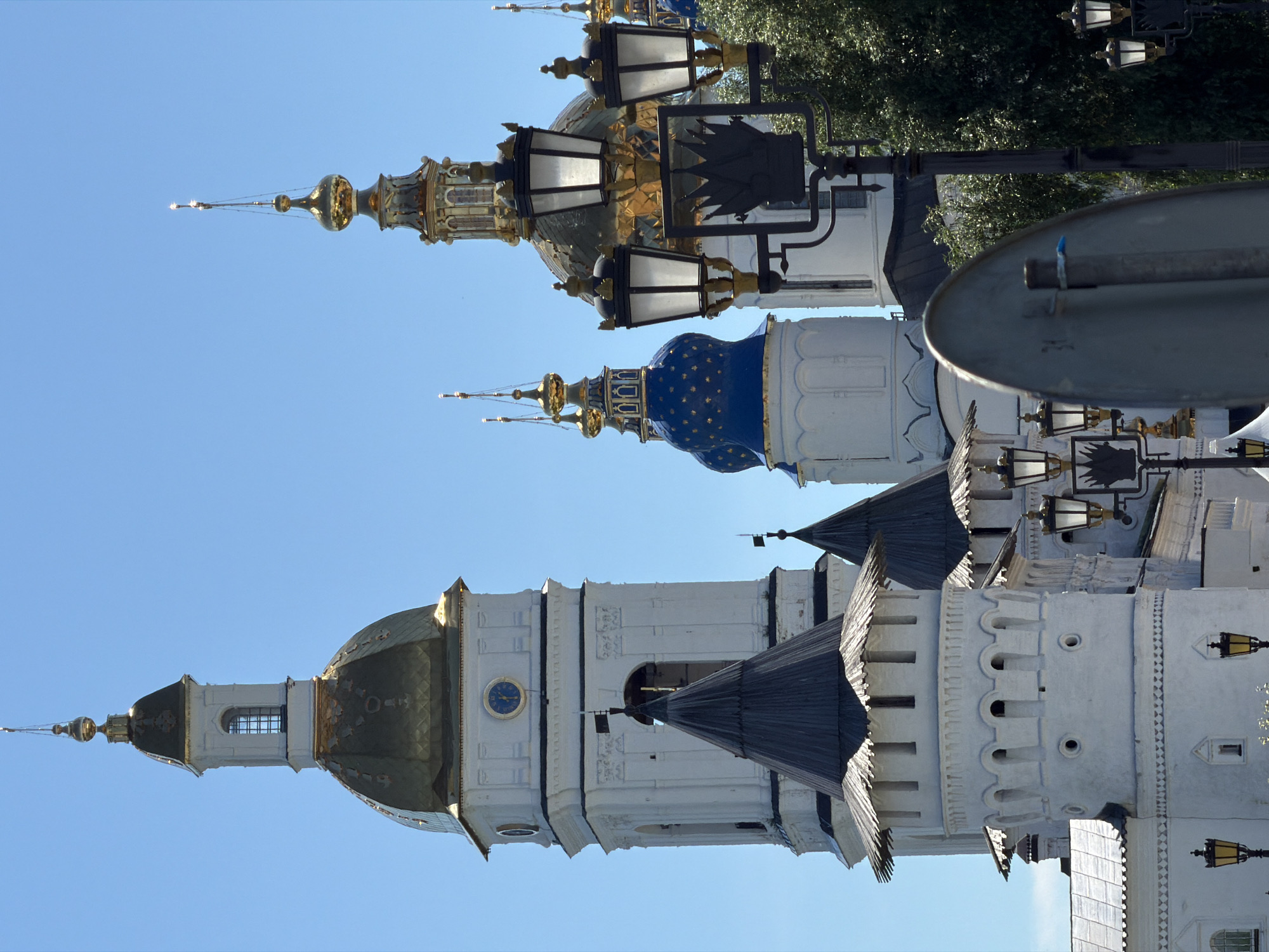 A vertical, eye-level, outdoor, close-up shot captures a bright, clear blue sky behind a cluster of white buildings topped with golden domes and spires. In the foreground, three ornate black street lamps with white glass globes are visible, with one in the center partially obscured by a silver, rounded sign. The buildings appear to be of historical or religious significance, with intricate architectural details such as arched windows, decorative trim, and what look like clock faces. One of the buildings on the left has a prominent tower with a golden dome and a smaller, white spire on top, while another building to its right features a dark, conical roof. Further to the right, a building with a large golden dome and several smaller golden spires are visible. The image is well-lit, suggesting it was taken on a sunny day.