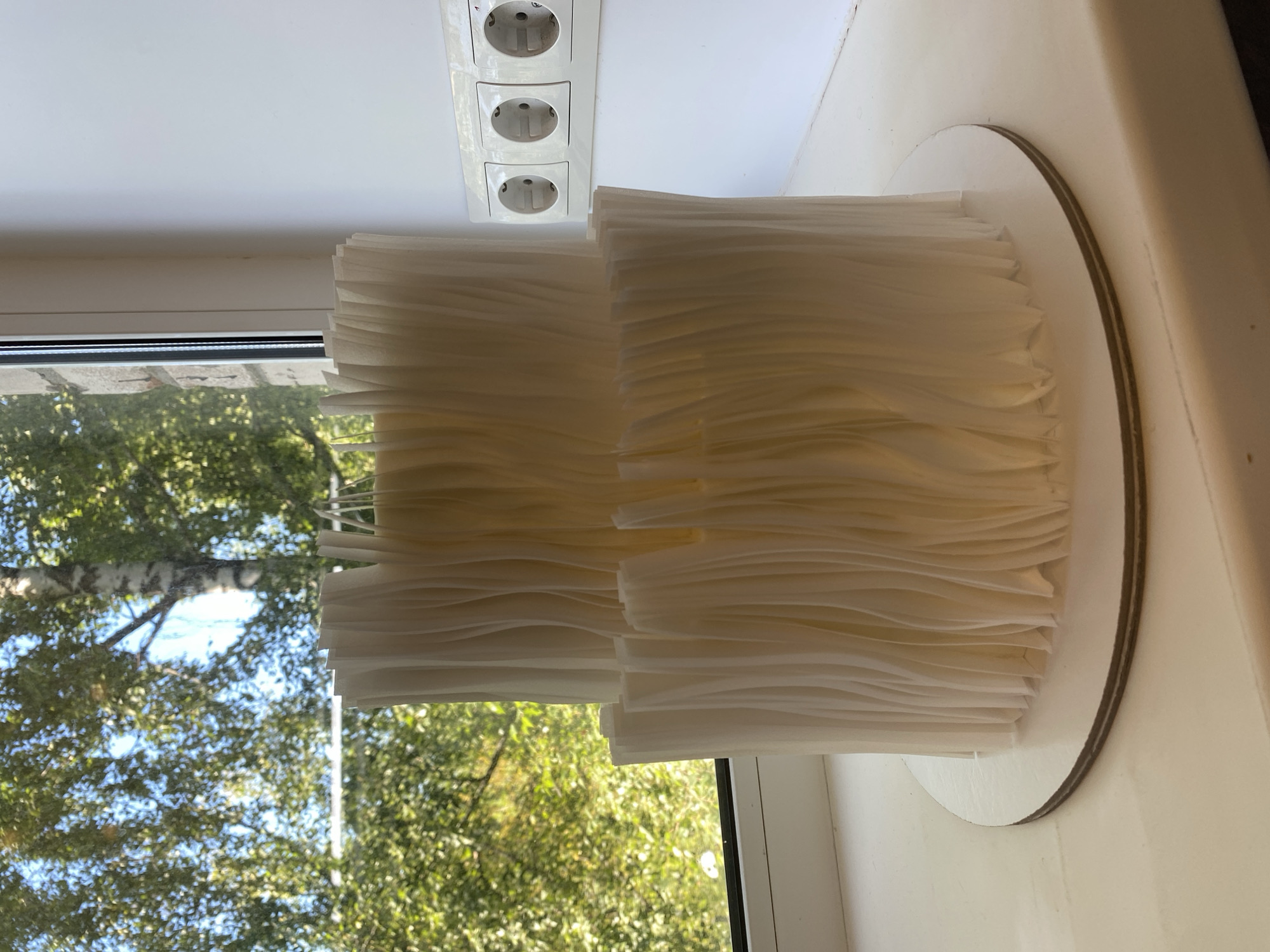 A two-tiered, cream-colored cake with a textured, pleated exterior sits on a white cake board. The cake is positioned on a white windowsill in front of a window that looks out onto trees and foliage. To the right of the cake, on the wall, are three white electrical outlets. The scene appears to be indoors, with natural light filtering through the window.
