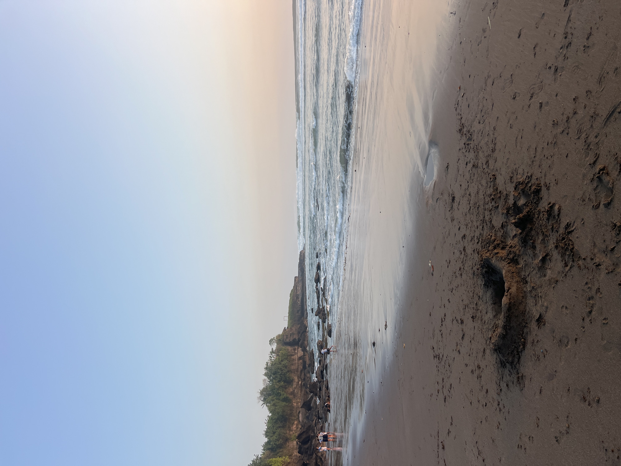 A wide, sandy beach in Kediri, Indonesia, is depicted with the ocean and a rocky cliff line. The sky is a clear, pale blue, transitioning to a soft, peachy hue near the horizon, indicating either sunrise or sunset. Gentle waves are rolling onto the shore.
In the foreground, dark, wet sand is visible, with a small, circular mound of sand, possibly a sandcastle, in the lower center of the frame. Scattered footprints are imprinted in the sand.
Further back, on the wet sand closer to the water, several individuals are present. Three people are standing together near the water's edge on the left side of the image. Another person is walking further down the beach. A few other individuals are scattered along the shoreline, some appearing to be children.
The background features a rocky cliff face covered with lush green trees and vegetation. The ocean stretches out to the horizon, with visible waves breaking. The overall scene is serene and suggests a leisurely day at the beach.