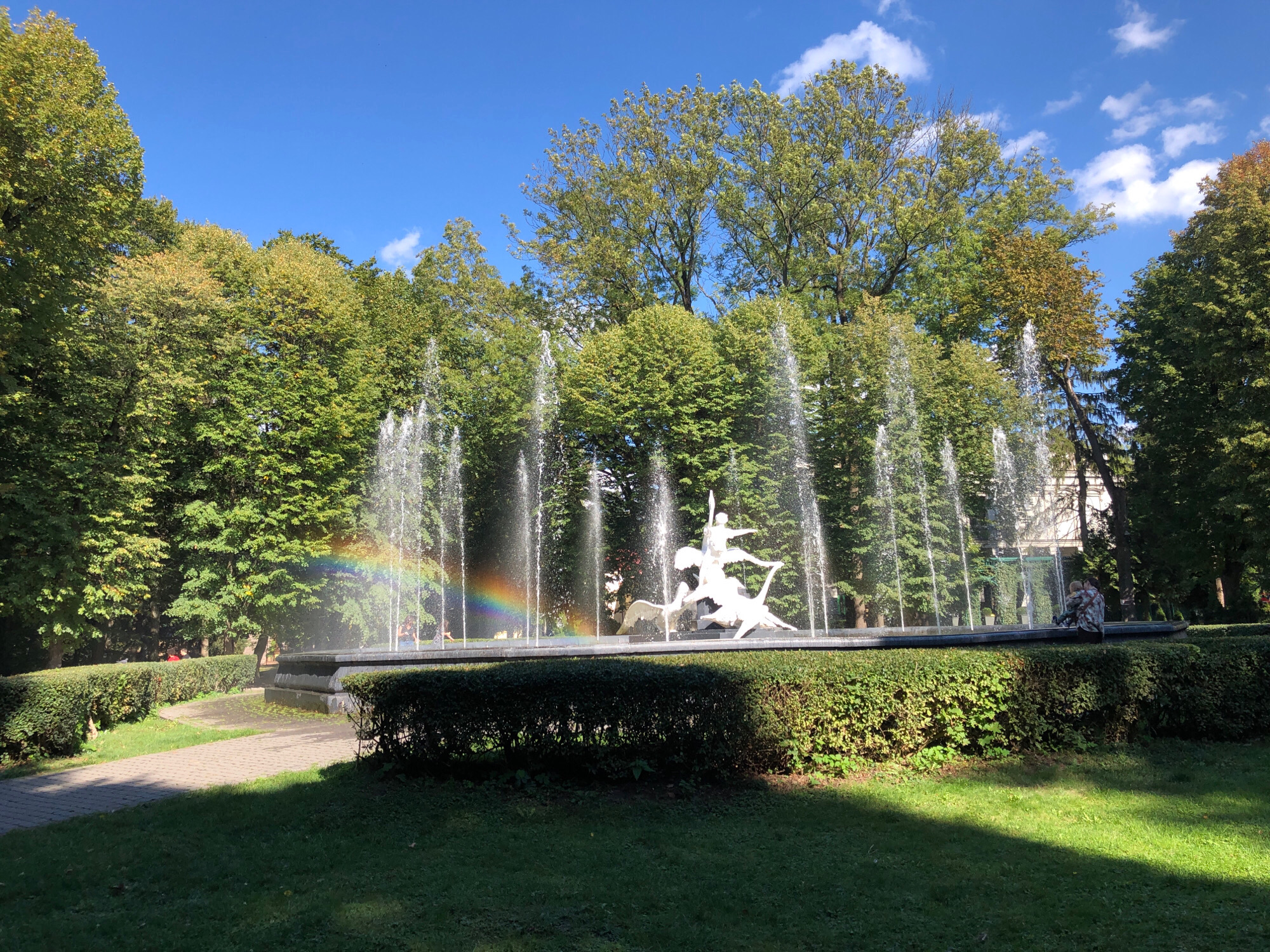 A wide shot captures a sunny day in Lviv, Ukraine, focused on a prominent fountain in a park. The fountain features a white sculpture of a human figure riding a horse-like creature, with water jets spraying upwards. To the left of the fountain, a rainbow is visible, created by the water spray. Surrounding the fountain is a well-maintained garden with manicured hedges and green lawns. In the background, lush green trees form a dense canopy, with a clear blue sky dotted with a few white clouds above. A paved pathway curves through the grassy area in the foreground, leading towards the fountain. On the right side of the fountain, two individuals are seated on the edge. One appears to be an adult, and the other a child.