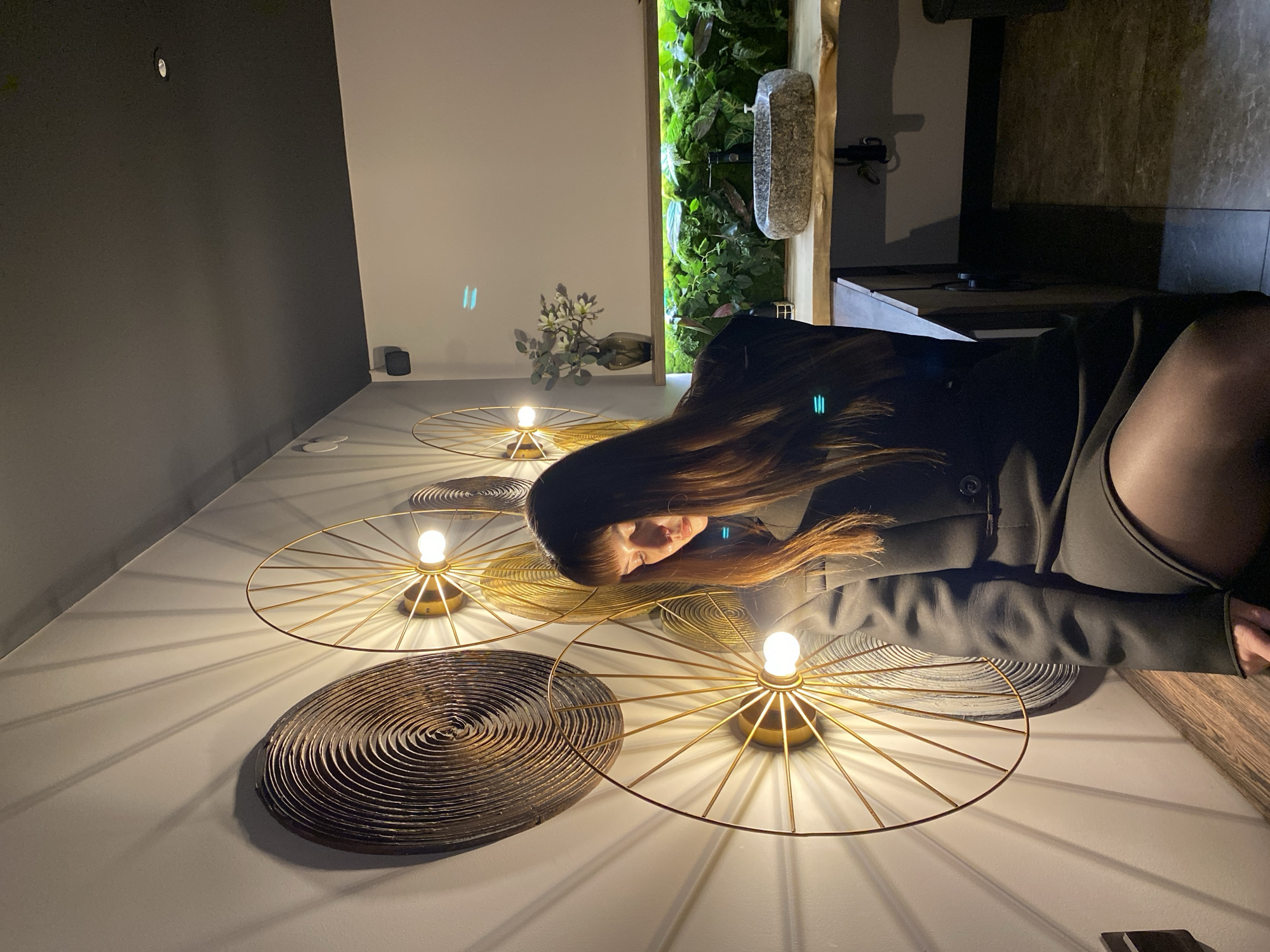 The image appears to be an interior shot of a room with a woman laying on a surface, likely a floor or table. There are multiple circular light fixtures with glowing bulbs arranged on the surface, casting light and shadows. The woman has long brown hair and is wearing a dark-colored jacket and dark tights. In the background, there is a wall divided into two sections - one is dark grey and the other is white. There is also a vertical arrangement of greenery with a decorative stone-like element. There is a vase with branches on a surface near the white part of the wall. The image does not contain enough information to determine a specific location or event, and I do not have any information about the city of Votkinsk, Russia.