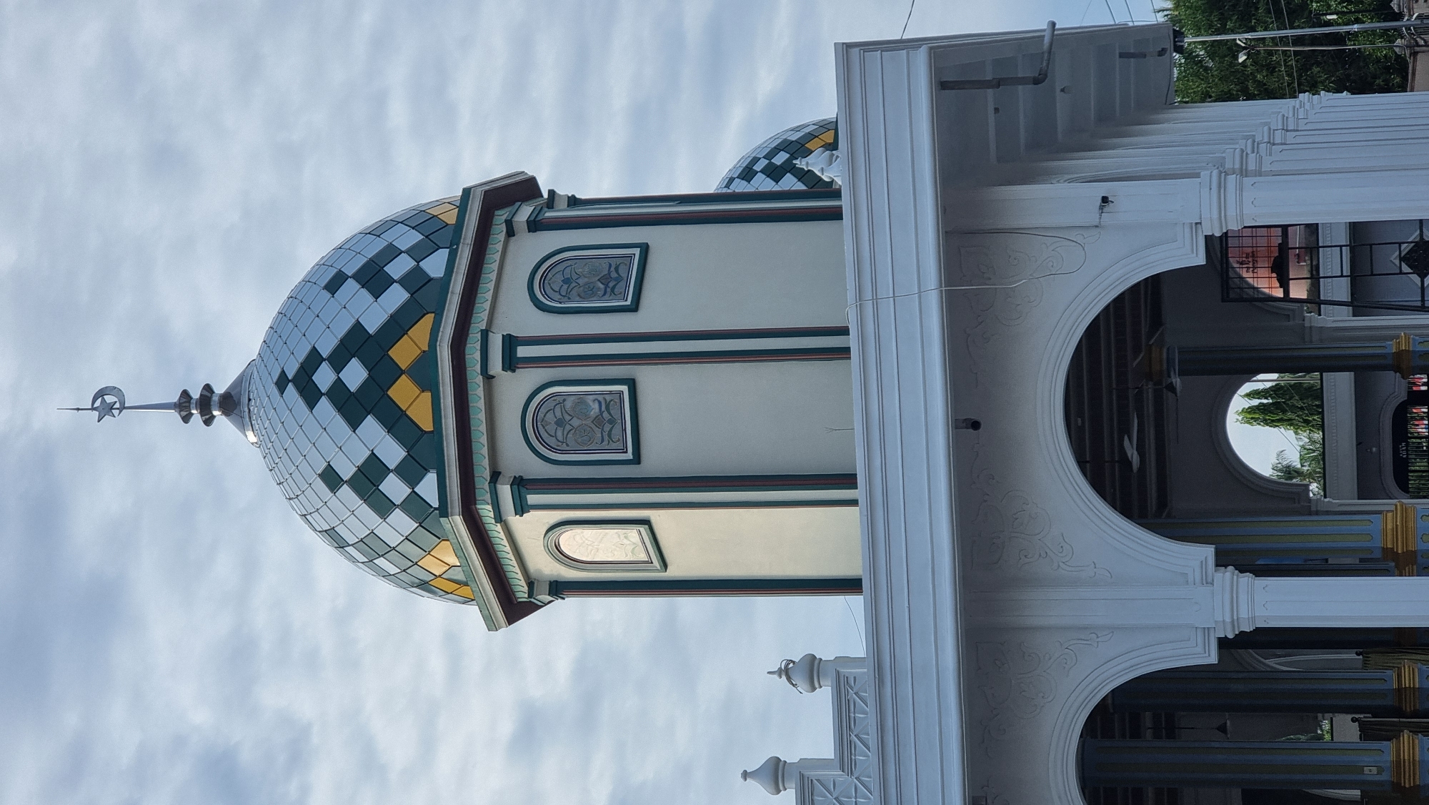 The image shows the exterior of a mosque in Meunasahlhok, Indonesia, under a cloudy sky. The central focus is a large, octagonal dome covered in a repeating pattern of diamond shapes in silver, green, and yellow. A silver crescent moon and star finial tops the dome. Below the dome, the light-colored wall features three arched windows with stained glass designs, flanked by vertical green and red trim. A smaller, similar dome is partially visible to the right. In the foreground, a white, ornate portico with multiple arches supported by columns leads into the mosque. Some of the columns are decorated with blue and yellow accents. The interior of the mosque is visible through the arches, with ceiling fans and lighting fixtures. Trees and a street light are visible in the background, indicating an urban or semi-urban setting. There are no people visible in the image.