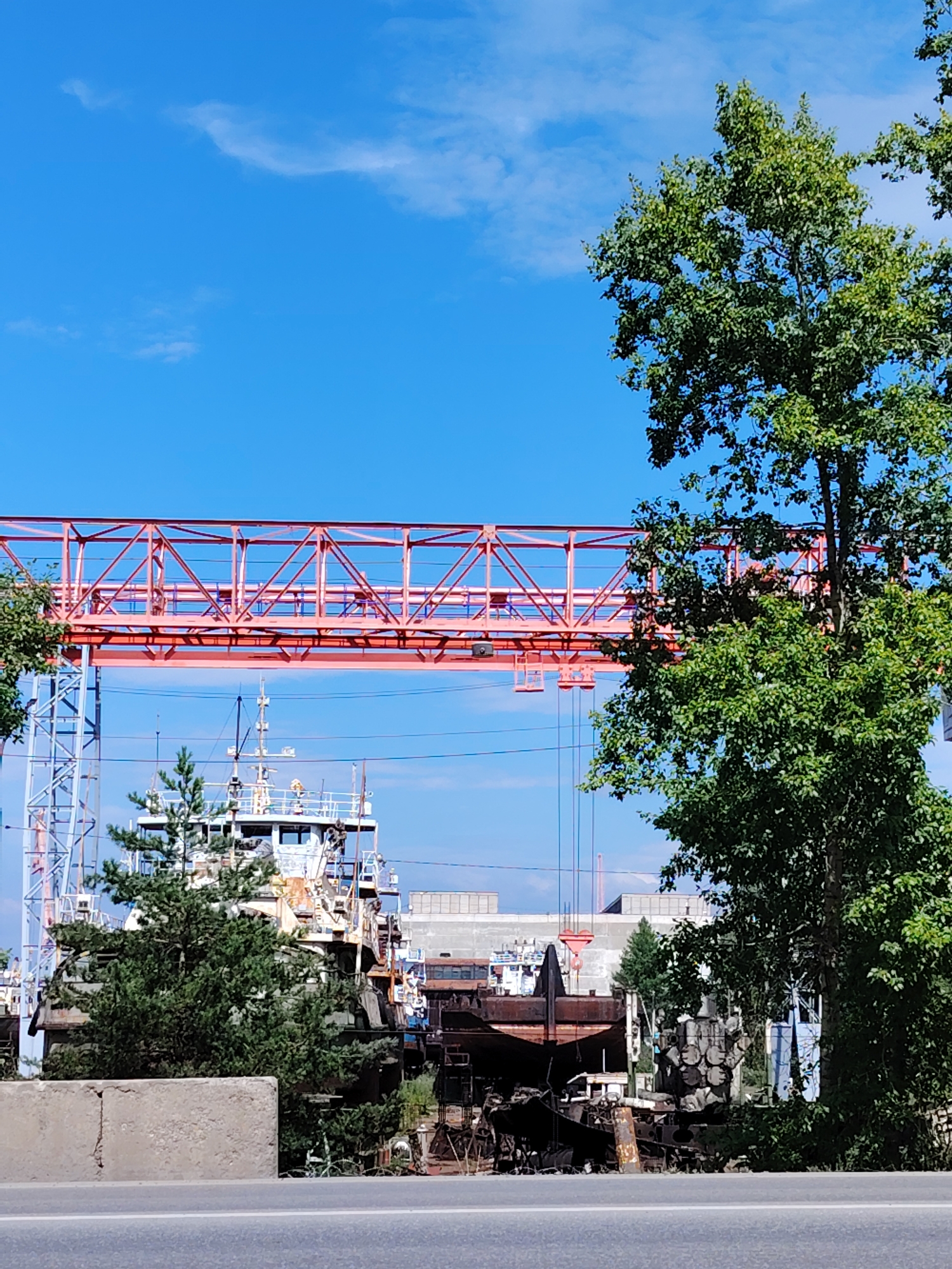 The image captures a view of a shipyard in the city of Chaikovsky, Russia. In the foreground, a grey concrete barrier separates a paved road from the shipyard's interior. Beyond the barrier, a large, rusty ship hulk is visible, partially obscured by green foliage. To the left, a portion of another vessel with a white superstructure and a green evergreen tree in front of it can be seen. A tall, orange metal gantry crane spans across the scene, positioned behind the ship. In the background, industrial buildings are visible under a clear blue sky with scattered white clouds. The overall impression is that of a working or decommissioned shipyard with a mixture of operational and derelict vessels and equipment. There are no people visible in the image.