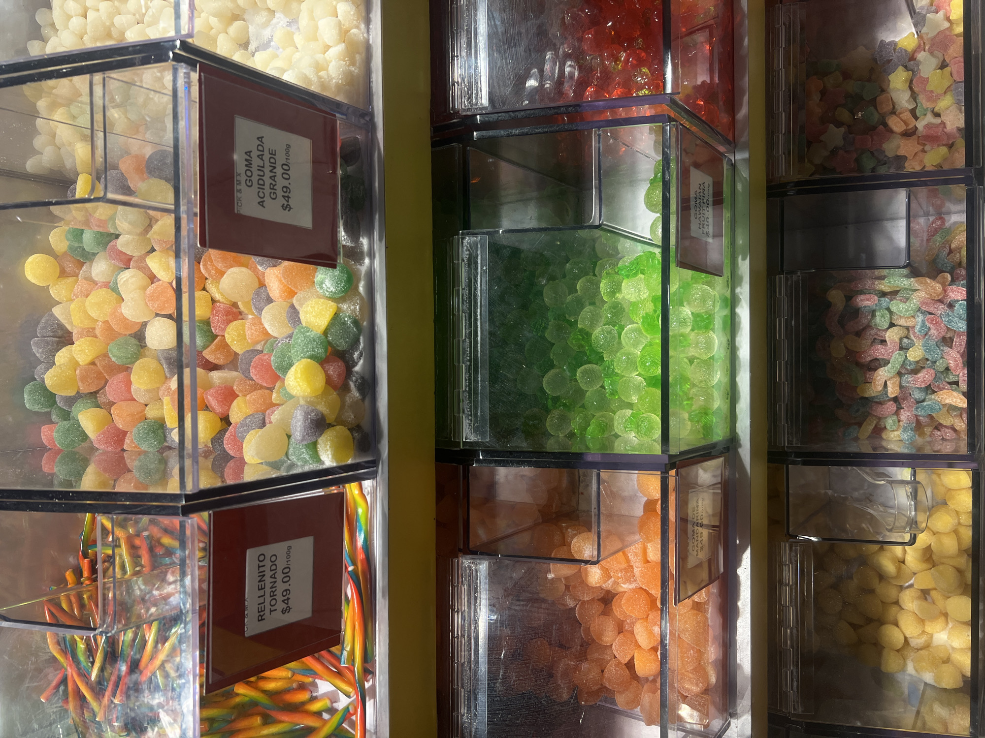 This image shows a vertical arrangement of clear, plastic candy dispensers, likely in a store in Mexico City, Mexico. The dispensers are filled with a variety of colorful candies, including white, and orange balls, green balls, red balls, worms and gummies. Labels are attached to the front of some dispensers, providing the name of the candy and price per 100g, which appears to be $49.00. One label reads "RELLENITO TORNADO" and another reads "GOMA ACIDULADA GRANDE." The setting is indoors, with a yellow-painted wall visible in the background.