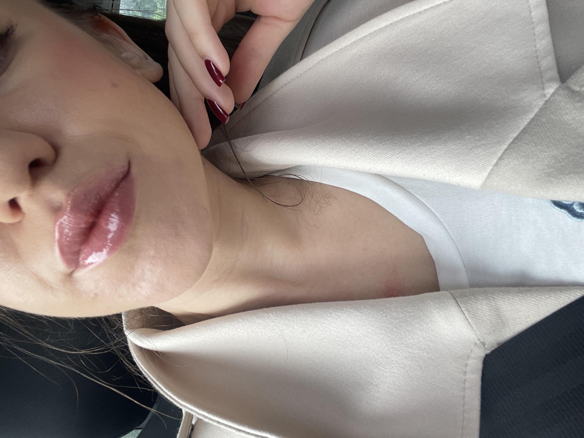 The image presents a close-up, diagonally oriented view of a person, likely taken in Sofia, Bulgaria. The subject's face is partially visible on the left, featuring the left side of their nose, cheek, and prominently, glossy, full lips tinted with a light pink or rose shade of lip gloss.
A hand, adorned with long nails painted in a deep red or burgundy polish, is positioned near the right side of the subject's face, possibly touching their ear or hair. Strands of dark hair are visible around the neck and shoulder area. The person is dressed in a light-colored, possibly beige or off-white, blazer or coat worn over a white t-shirt. The texture of the outer garment is discernible.
The background is softly blurred, with hints of a dark surface or garment on the right and a suggestion of an outdoor scene or window in the upper left, implying the photograph was taken indoors or within a vehicle. The lighting is soft and even, emphasizing the subject's features and manicure. No visible text is present in the image. The overall impression is a posed, close-up portrait with a focus on makeup and accessories.