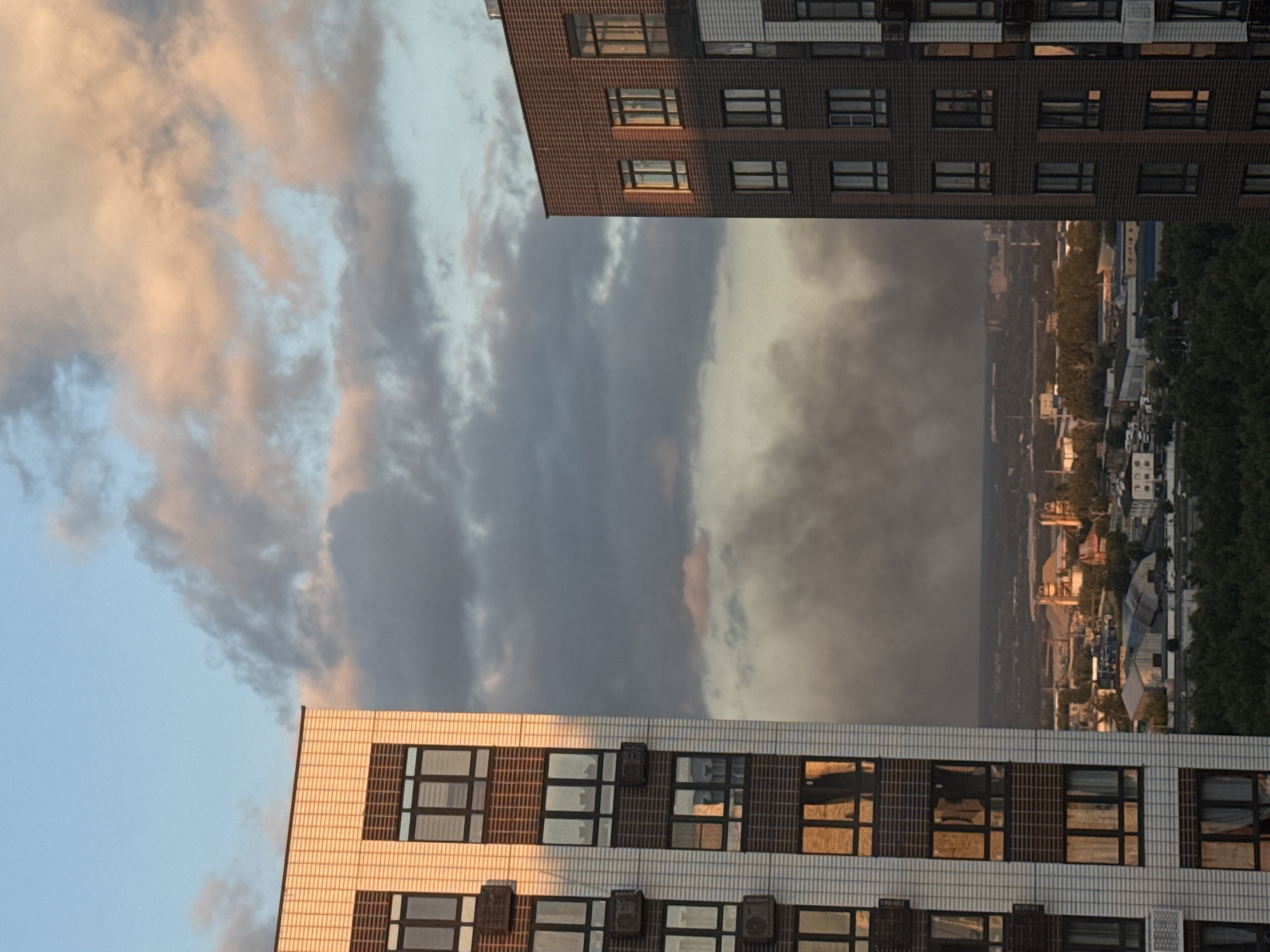 The image is an outdoor, eye-level shot of a city skyline in Lyubertsy, Russia, during what appears to be either sunrise or sunset. Two modern apartment buildings dominate the foreground, one on the left with a white brick facade and the other on the right with a dark brick facade. Both buildings feature multiple windows and air conditioning units.
In the midground, a dense urban landscape stretches across the horizon, dotted with various structures including what appear to be industrial buildings, warehouses, and smaller residential buildings. A significant plume of dark smoke is visible in the distance, rising into the sky and obscuring a portion of the horizon, suggesting a potential fire or industrial activity.
The background consists of a dramatic sky filled with clouds. The clouds are a mix of dark gray and lighter pink and orange hues, illuminated by the low-angle sunlight, creating a striking contrast. The overall impression is one of an urban environment under a dynamic sky, with the presence of smoke adding a somber element to the scene. No people are visible in the frame.