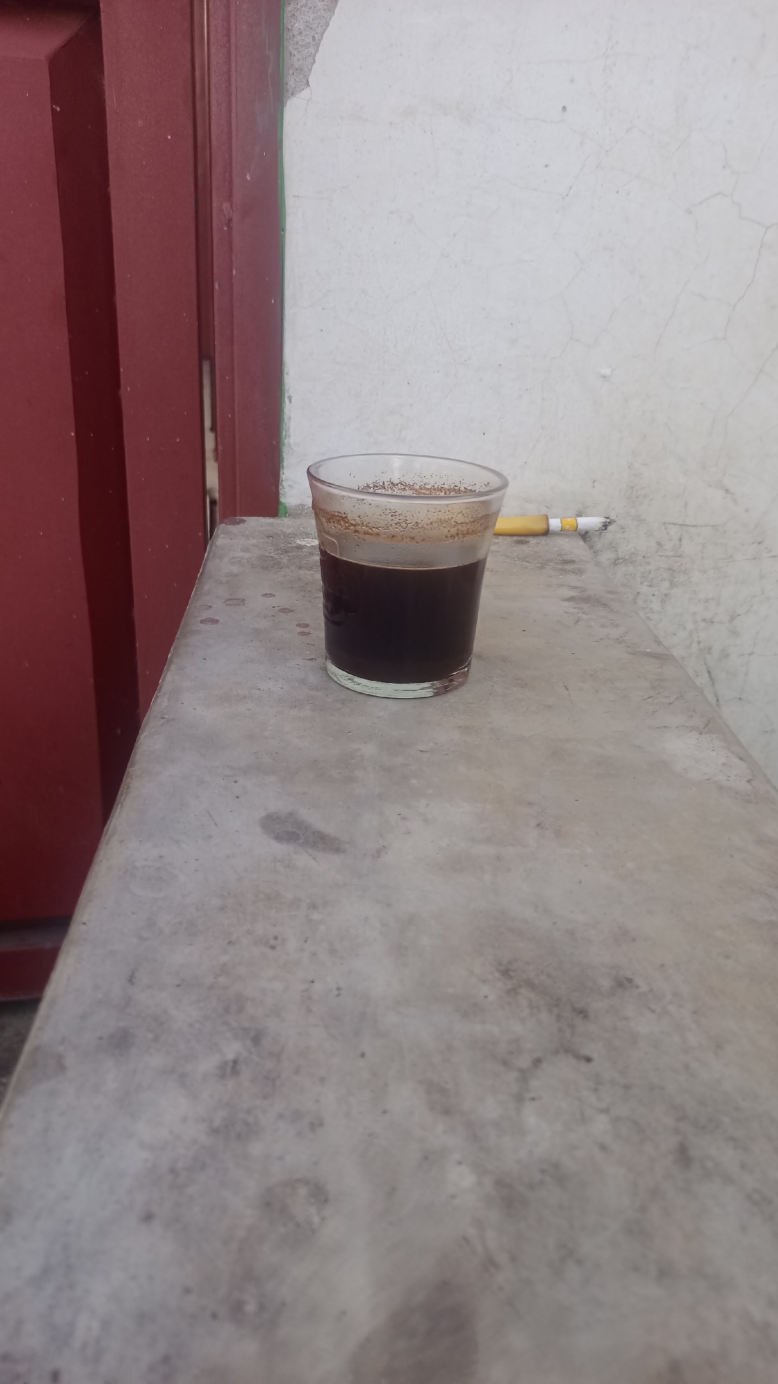 In Pakemitan, Indonesia, a clear glass of dark coffee sits on a concrete surface. The coffee has a thin layer of foam and sediment at the top. To the right of the glass, a partially smoked cigarette with a yellow filter is visible. The background consists of a textured white wall with visible cracks and a portion of a red door or paneling on the left. The lighting suggests an outdoor or well-lit interior space.