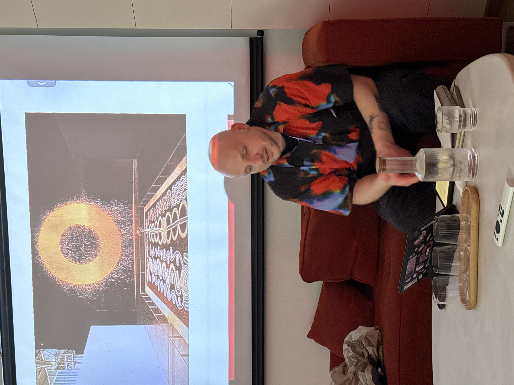 An indoor scene depicts a man sitting on a reddish-brown sofa, facing a large projection screen that displays abstract art. The man is bald with a short beard, wearing a vibrant, patterned shirt in black, red, and blue, and black pants. Tattoos are visible on his left arm. He appears calm, with a neutral to slightly smiling expression.
The projection screen, dominating the left side of the frame, showcases an image with a large, glowing golden ring or halo, its edges appearing speckled, against a dark background. To the right of this ring, there is an intricate pattern of white, wavy lines and shapes on a darker, possibly brown or black, surface. A small, left-pointing arrow icon is visible at the top left corner of the projected content.
To the right of the man, a white table holds several clear glass tumblers, a clear glass bottle with a cork stopper, and a wooden tray. A white remote control and a tablet or laptop with a decorative cover are also on the table. A crumpled bag or paper is visible on the floor next to the sofa. The setting suggests a casual gathering, presentation, or viewing event. The image provides no specific time of day or weather cues, but the indoor lighting implies it could be any time. The location context is Moscow, Russia.