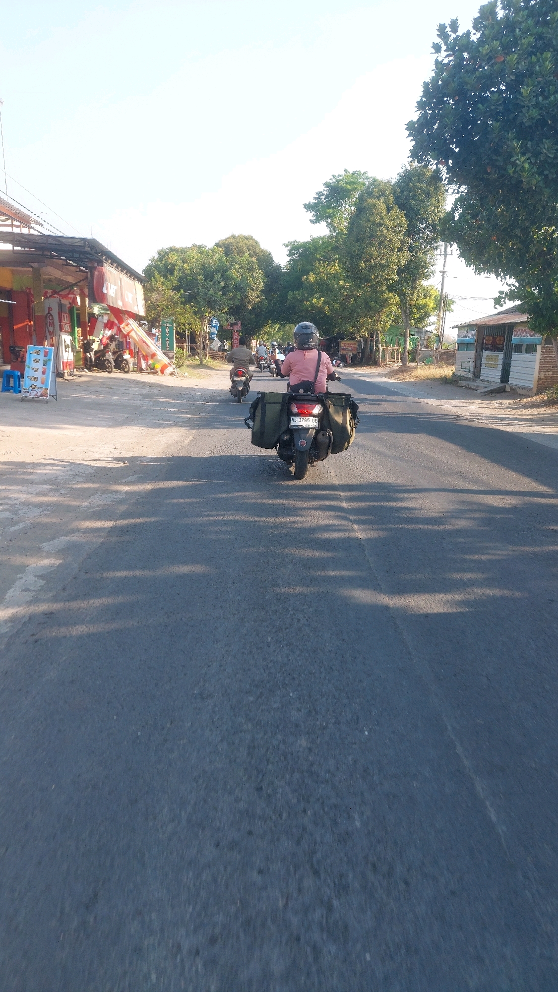 The image shows a person riding a motorcycle down a paved road in the city of Ngrupit, Indonesia. The motorcycle has two large olive-green bags attached to the rear, one on each side of the passenger seat. The rider is wearing a pink shirt and a black helmet. The road is flanked by trees and a few buildings, including a storefront on the left side of the road with various signs and posters. Several other motorcycles are visible on the road ahead of the main subject, some with riders and some without. The sky is clear and bright, suggesting it is daytime. The sunlight casts shadows across the road, indicating a low sun angle, possibly early morning or late afternoon.
