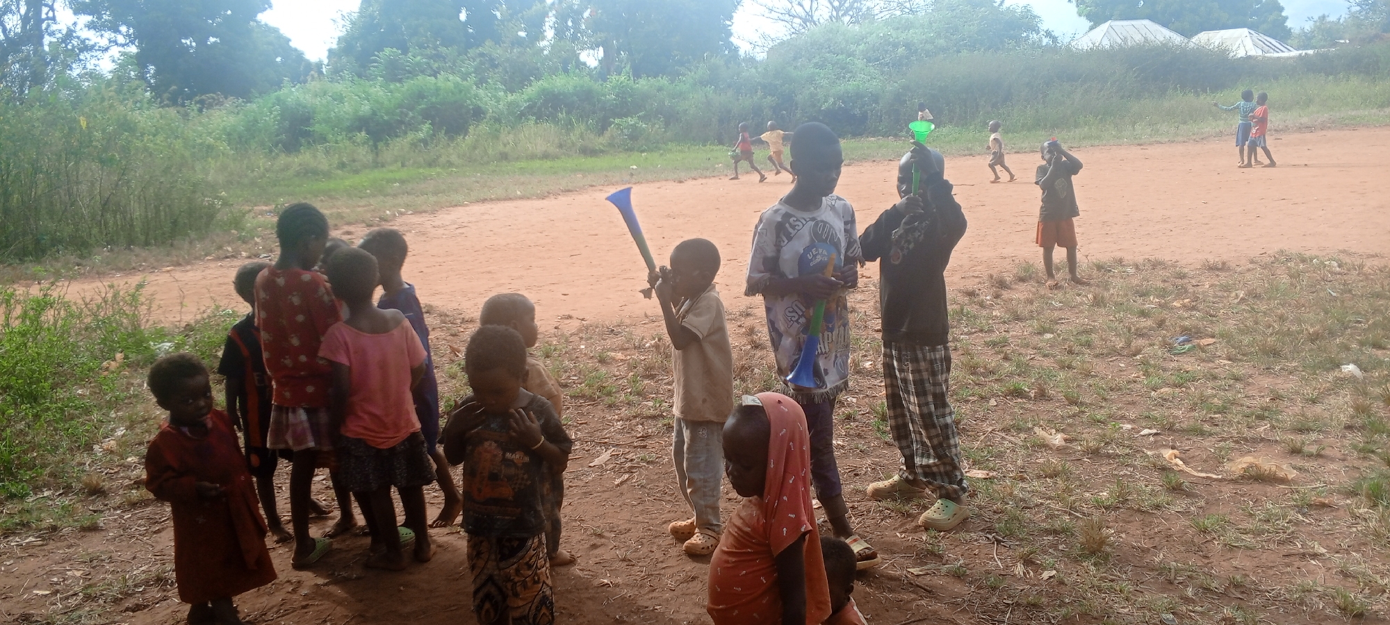 The image shows a group of children in Ngalula, Democratic Republic of the Congo. Some children are standing in a group on the left side of the image, while others are playing in the background on a dirt field. Some children are holding and playing with vuvuzelas. The environment appears to be rural, with trees and vegetation surrounding the dirt field. There are a few buildings visible in the distance.