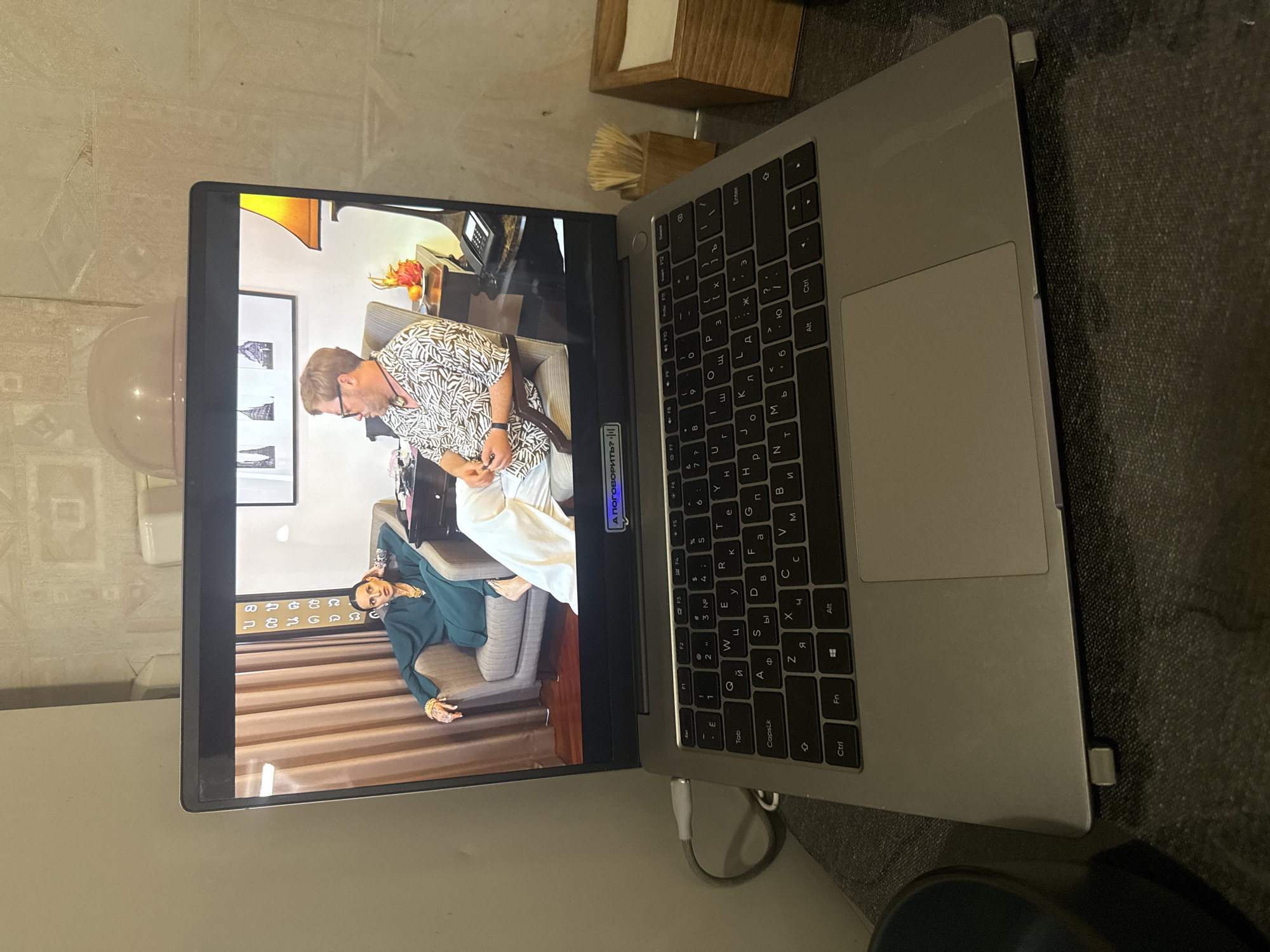 A laptop is open and placed on a dark textured surface. The laptop screen displays a video of two individuals sitting in chairs. On the left, a woman in a dark green dress is seated, leaning back with her head resting on her hand, wearing jewelry. On the right, a man with glasses and a patterned shirt is also seated. The background of the video shows a room with curtains and a lamp. Below the screen, the laptop keyboard is visible with Cyrillic characters on the keys. A USB cable is plugged into the side of the laptop. To the right of the laptop, a wooden container with toothpicks and a small wooden box with tissues are present. The overall scene suggests an indoor setting. The image is taken from a slightly elevated angle, looking down at the laptop and its immediate surroundings. The laptop itself appears to be on a desk or table.