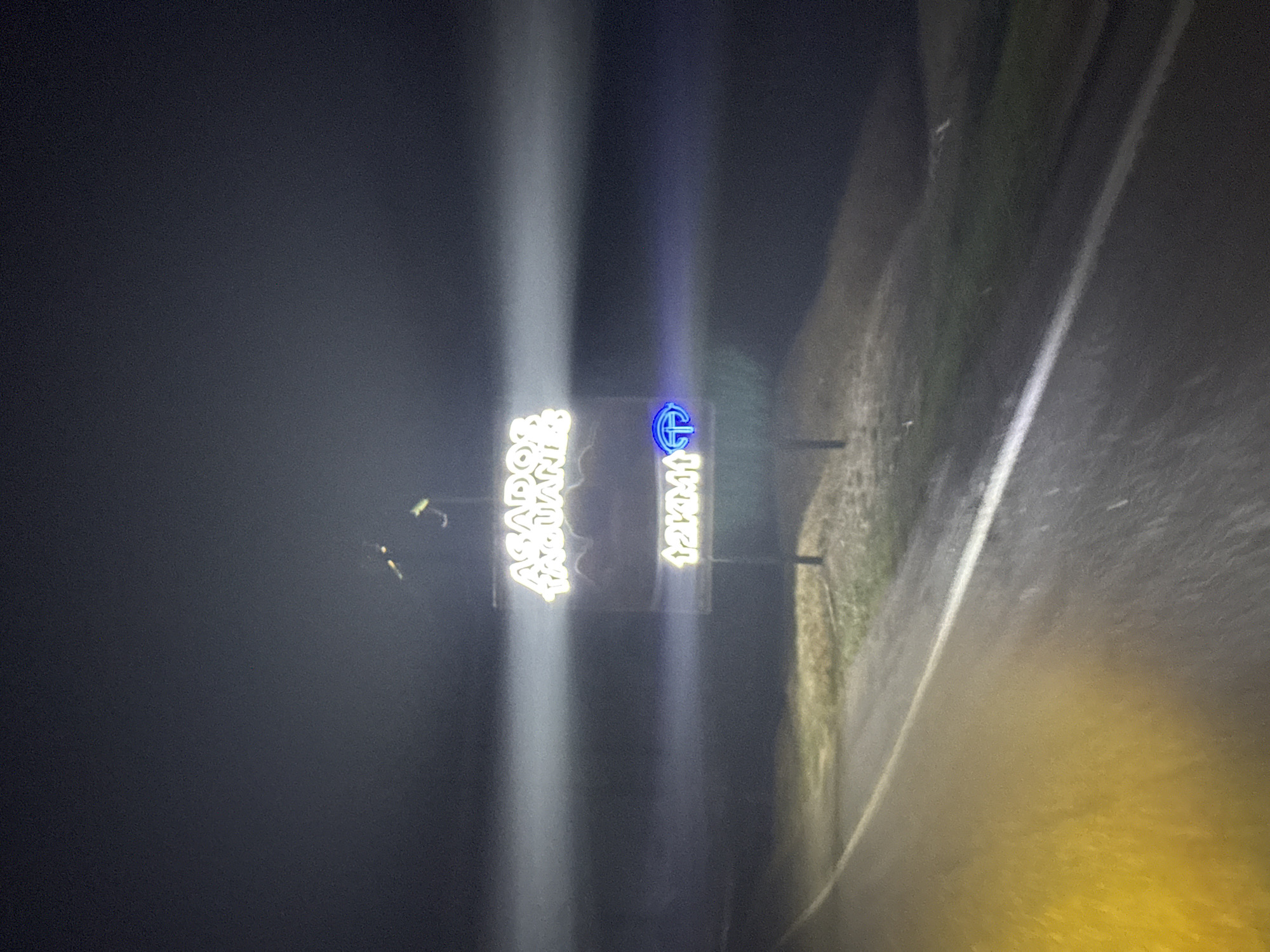 The image depicts a nighttime scene along a road in Tinaquillo, Venezuela. A sign on the left reads "ASADOR PAGUAN". Another sign to its right displays "12 KM", accompanied by an emblem. The road is illuminated by vehicle headlights.