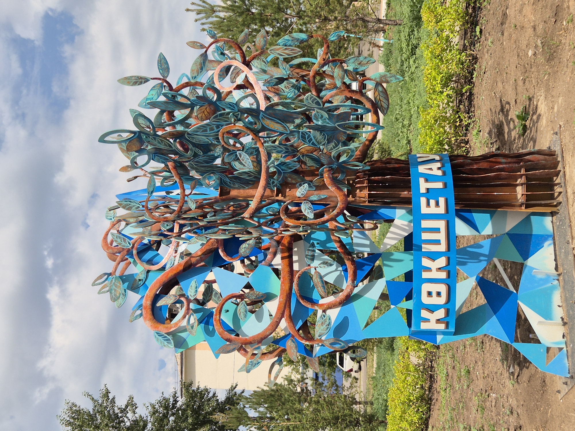 An outdoor metal sculpture resembling a tree with blue geometric shapes and brown, curling branches adorned with teal leaves is situated in a landscaped area. White text in a blue rectangular sign reads "ҚОКШЕТАУ" in the foreground. The sculpture is set against a backdrop of green bushes, grass, trees, and a partially visible building. The sky above is partly cloudy. The location appears to be a public space in the city of Kokshetau, Kazakhstan, as indicated by the text on the sign. There are no people or animals visible in the image.