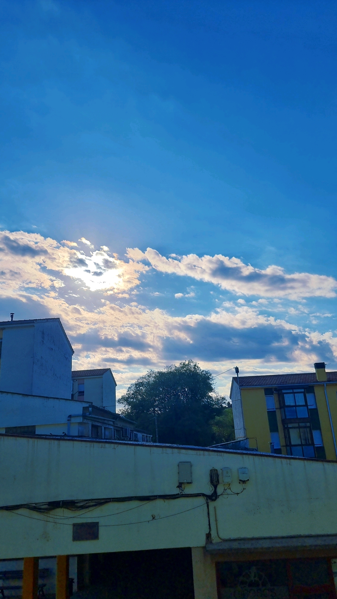 The image depicts a sky with scattered white and grey clouds, partially obscuring the sun. Below the sky are several buildings. In the foreground, a light yellow building with electrical conduits and boxes attached to its exterior wall runs horizontally across the frame. Behind this, to the left, is a white building with a visible roofline. Further back, a large green tree is situated between buildings. To the right of the tree is a yellow building with multiple windows. The scene appears to be an urban or suburban environment, likely in Sant Joan Les Fonts, Spain.