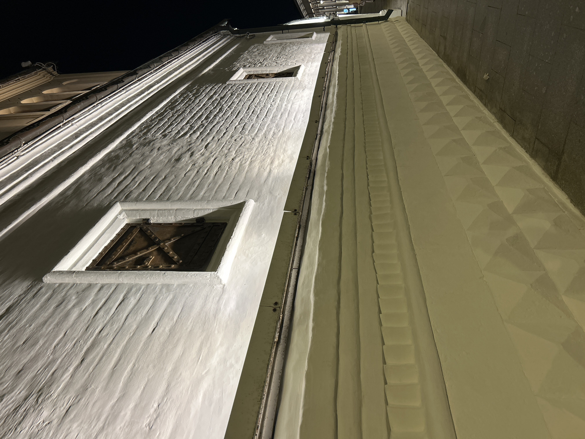 This is a nighttime, ground-level, eye-level, medium shot of a textured white brick building facade with two small, dark, barred windows. The building is illuminated by spotlights from below, creating dramatic shadows and highlighting the rough texture of the bricks. The lower part of the building features a lighter, cream-colored section with horizontal molding and a geometric, diamond-shaped pattern at the very bottom. A dark, paved walkway runs along the base of the building, and a small piece of white debris is visible on the ground. The image is captured at an angle, with the building receding into the background. The style of the building and its location suggests it is likely in the historical center of Moscow, Russia, possibly part of a monastery or an older administrative building. The scene is devoid of people.