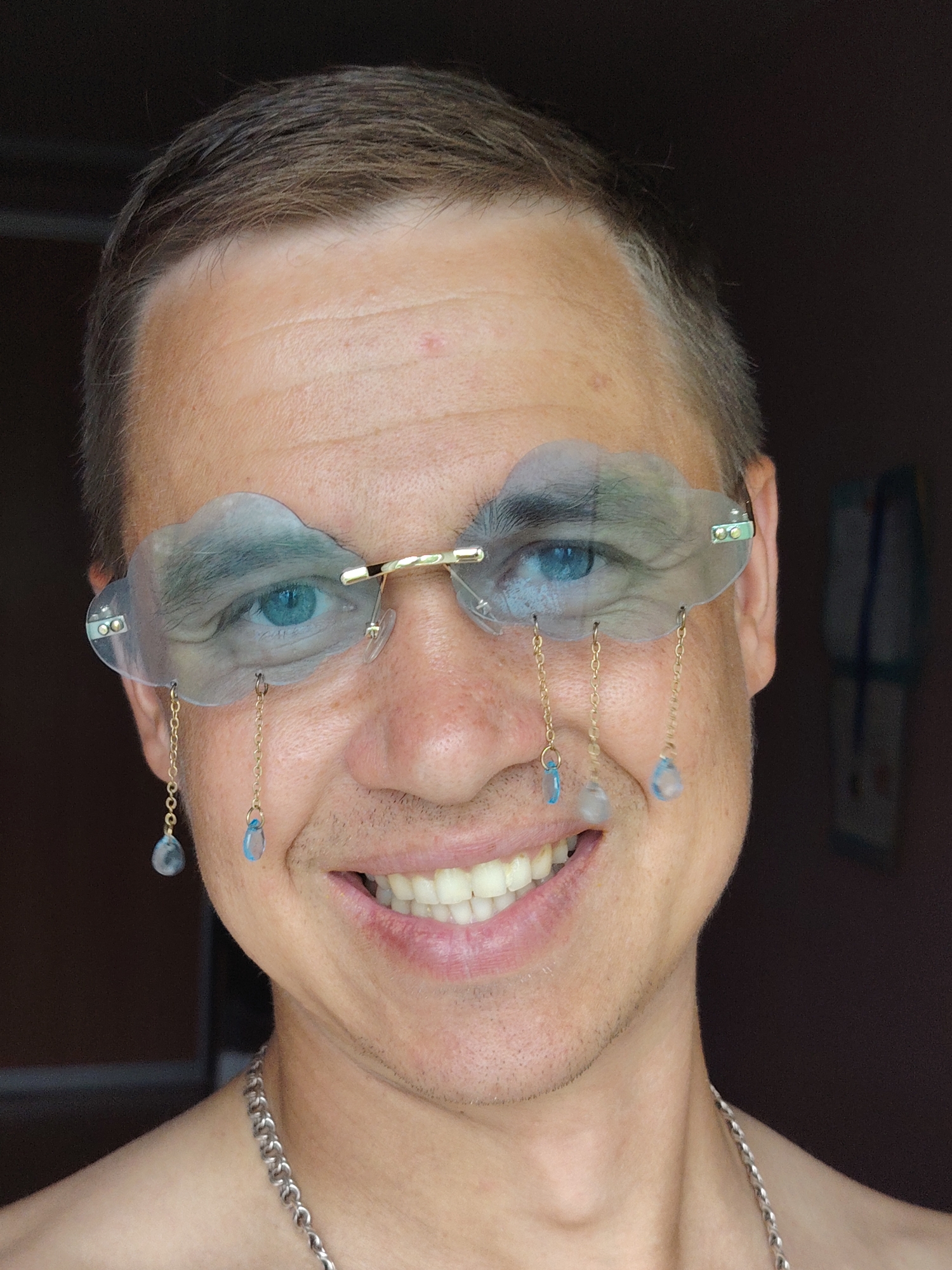 A frontal, close-up portrait of an adult male subject in the city of Chaikovsky, Russia. The man has short, light brown hair, fair skin with some minor facial stubble around the mouth and chin, and blue eyes. He is smiling broadly, revealing his upper and some lower teeth.
He is wearing a pair of novelty glasses with transparent, cloud-shaped lenses that are slightly frosted. The glasses feature a gold-toned metal bridge and temple arms. Attached to the lower edge of each cloud lens are two delicate gold-toned chains, each terminating in a translucent, light blue, teardrop-shaped bead, resembling raindrops. A total of four such chains and beads are visible. A silver-toned curb chain necklace is worn around his neck, resting on his collarbone. His upper torso appears bare.
The background is dark, out of focus, and indistinct, suggesting an indoor setting. A faint, light-colored vertical element is partially visible on the right side of the frame. The lighting is direct, illuminating the subject's face and upper chest.