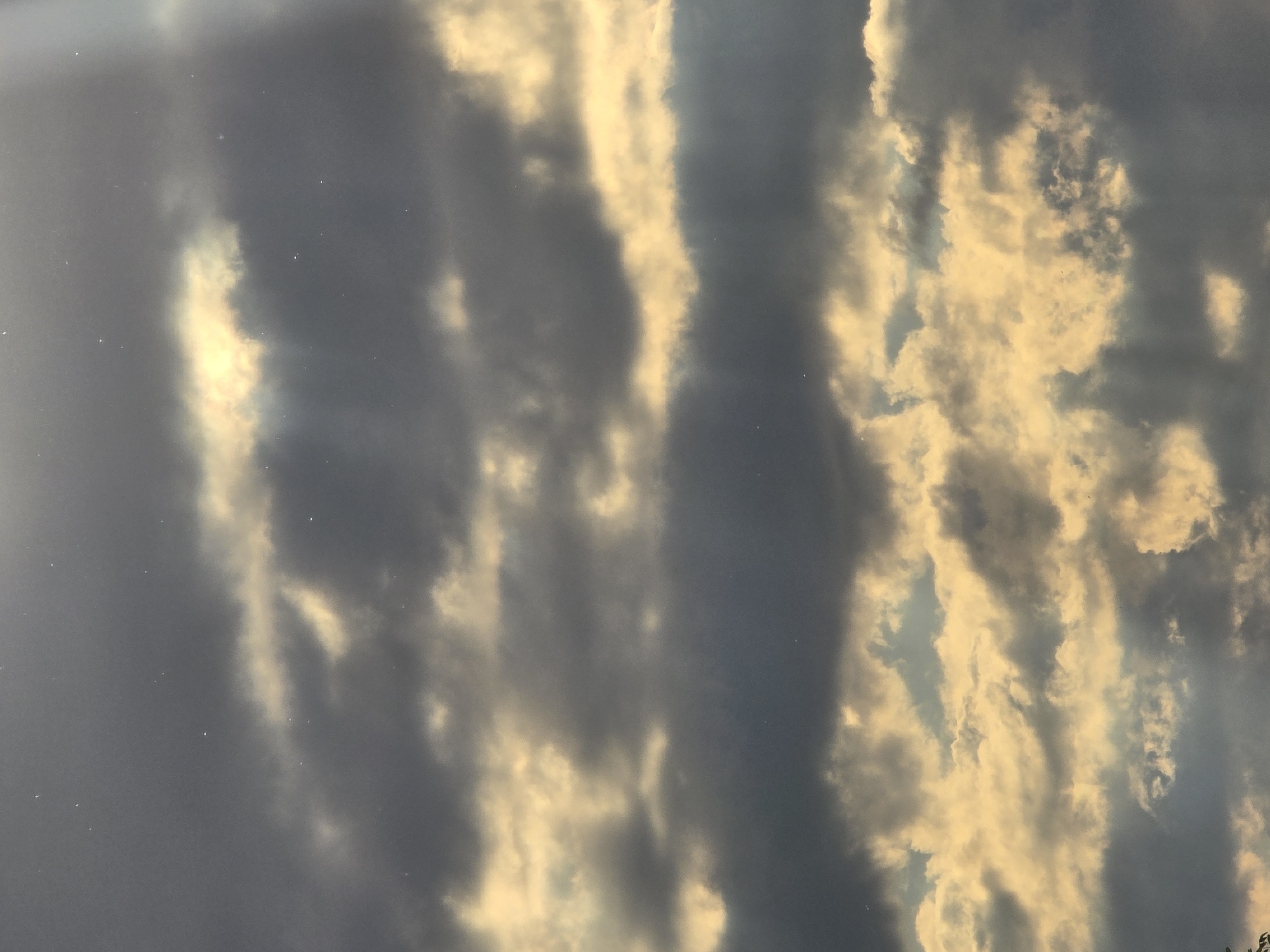 The image displays a cloudy sky, with alternating streaks of dark, gray clouds and bright, golden-lit clouds. The scene seems to be taken during sunrise or sunset, given the warm tone illuminating the clouds. The location is Unknown, Unknown.