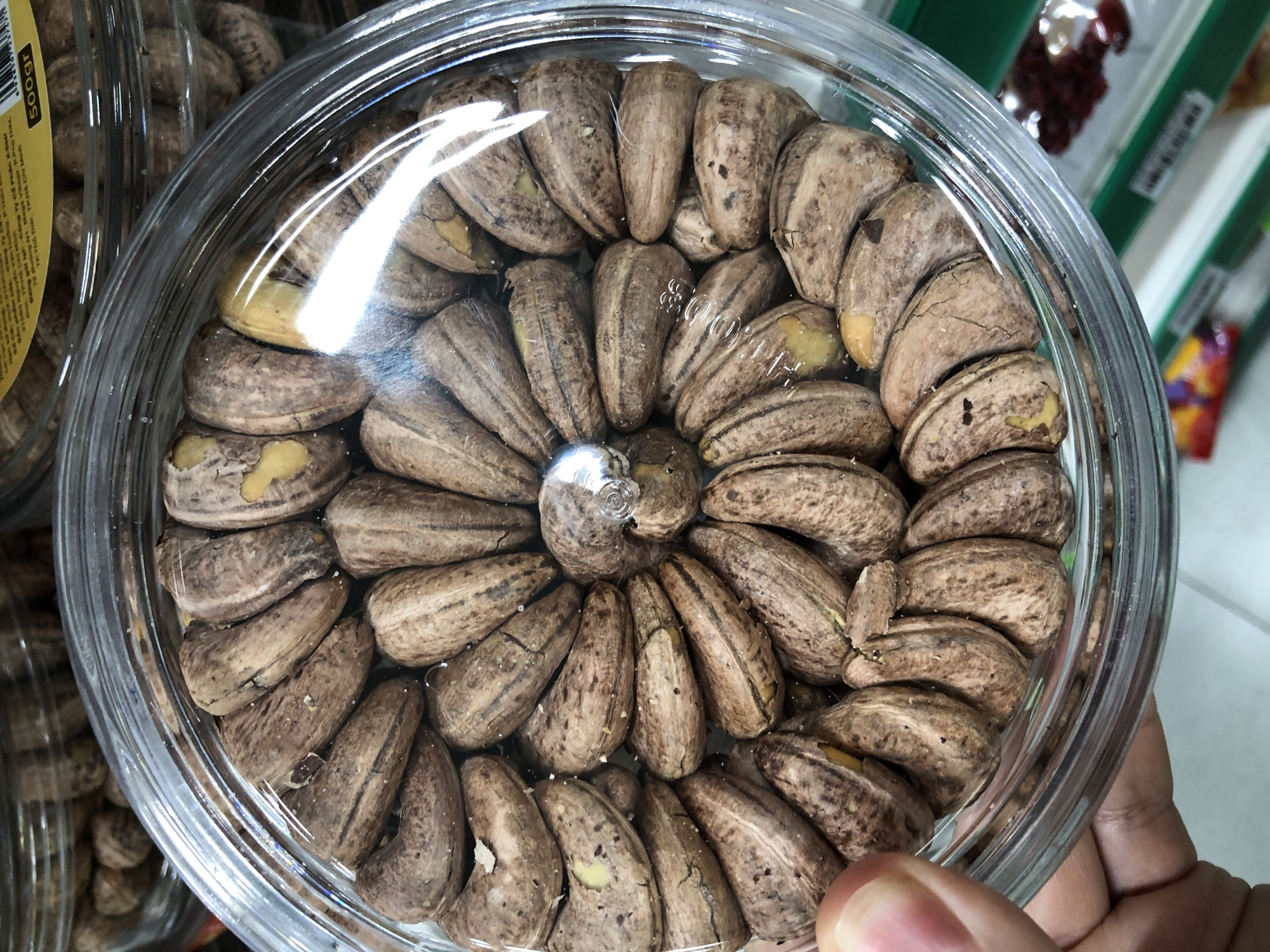 This is a close-up, top-down shot of a clear plastic container filled with cashew nuts. The container is round with a ridged lid, and the nuts are arranged in a circular pattern within it, filling the container to the brim. The cashews have their shells on, which are a mottled brown and tan color with some lighter yellow or cream-colored patches. Many of the shells show natural cracks and imperfections. The surface of the nuts is slightly textured.
In the background, partially visible through the clear plastic of the main container and out of focus, are other containers of what appear to be more nuts or similar food items. There are also glimpses of shelves and possibly other products in what looks like a retail environment, with some colorful packaging visible at the bottom edge. The lighting creates some glare on the plastic lid and the surfaces of the nuts. A person's thumb and part of their hand are visible in the lower left corner, resting on the rim of the container.
There is no visible information in the image that would indicate the location in Đà Nẵng, Vietnam, or any specific urban setting. The scene is purely focused on the product inside the container.