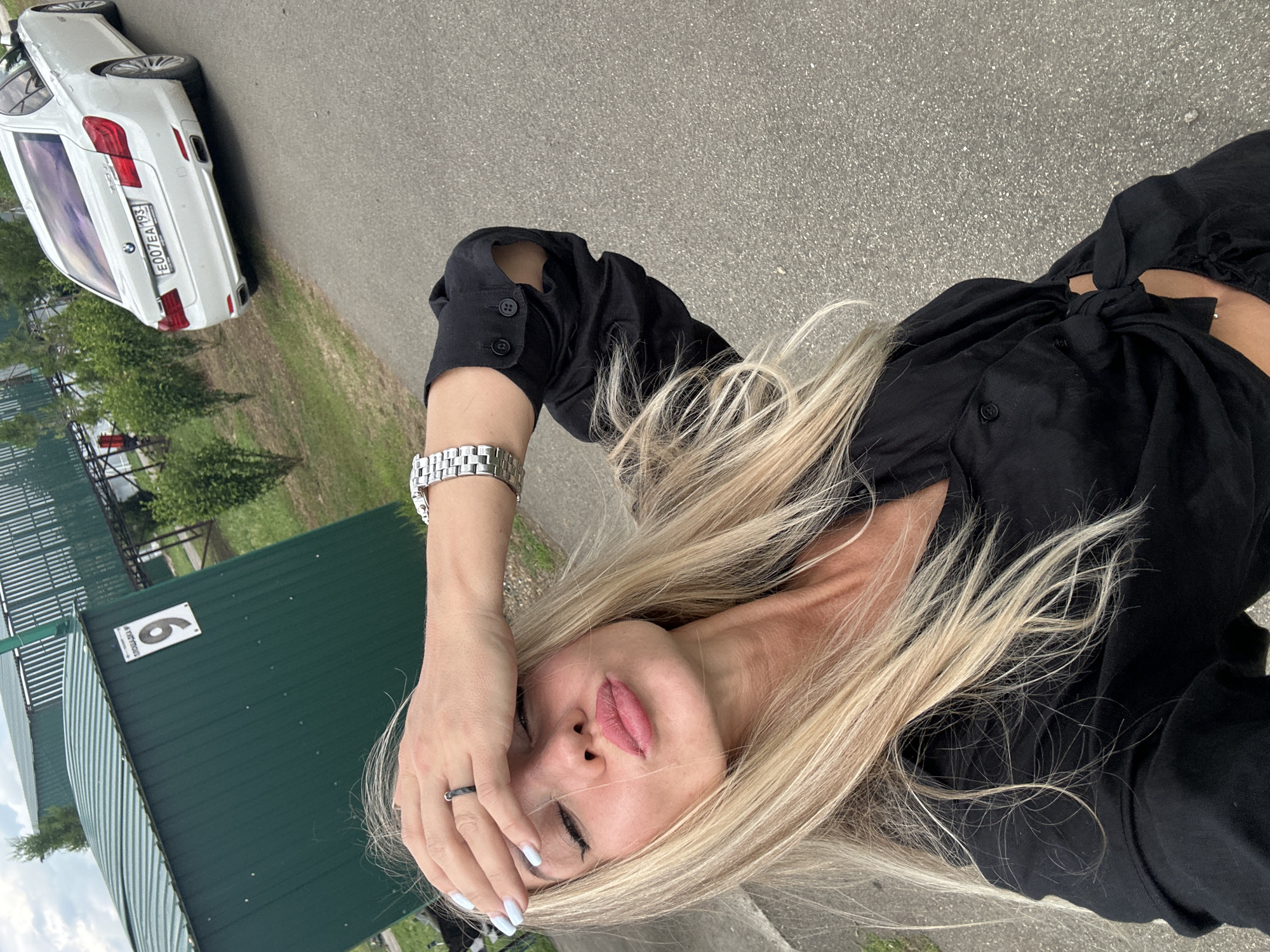 A woman with blonde hair is lying on the ground, holding her hand to her forehead. She is wearing a black shirt and a watch on her left wrist. Her eyes are closed, and her lips are slightly parted. A white car is parked in the background, and a green building with the number "6" on it is also visible. The scene appears to be outdoors, possibly in a park or recreational area. The weather seems to be cloudy. The text "E007EA 193" is visible on the license plate of the car. The overall mood of the image is one of relaxation or perhaps weariness.