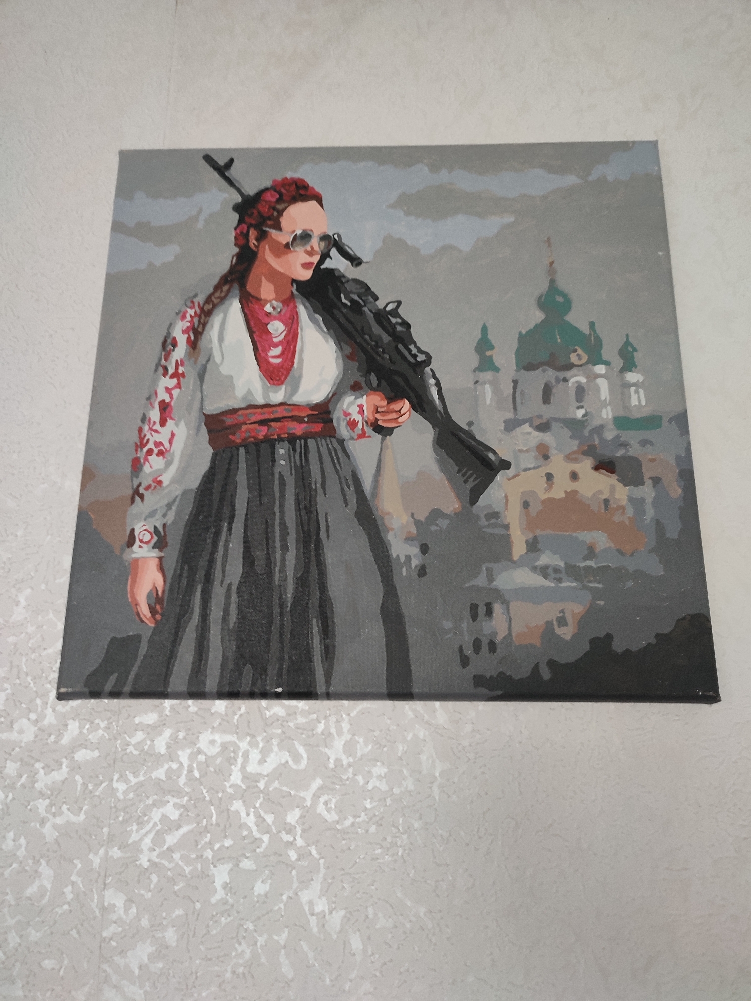 A painting depicts a woman in traditional Ukrainian clothing, a white embroidered shirt with red floral patterns and a long grey skirt, holding a rifle over her shoulder. She wears sunglasses and her hair is braided with red ribbons. In the background, a cityscape with domed buildings, possibly churches, is rendered in muted tones. The painting is displayed on a textured wall. There is no indication of the specific location within the city of Yaresky, Ukraine.