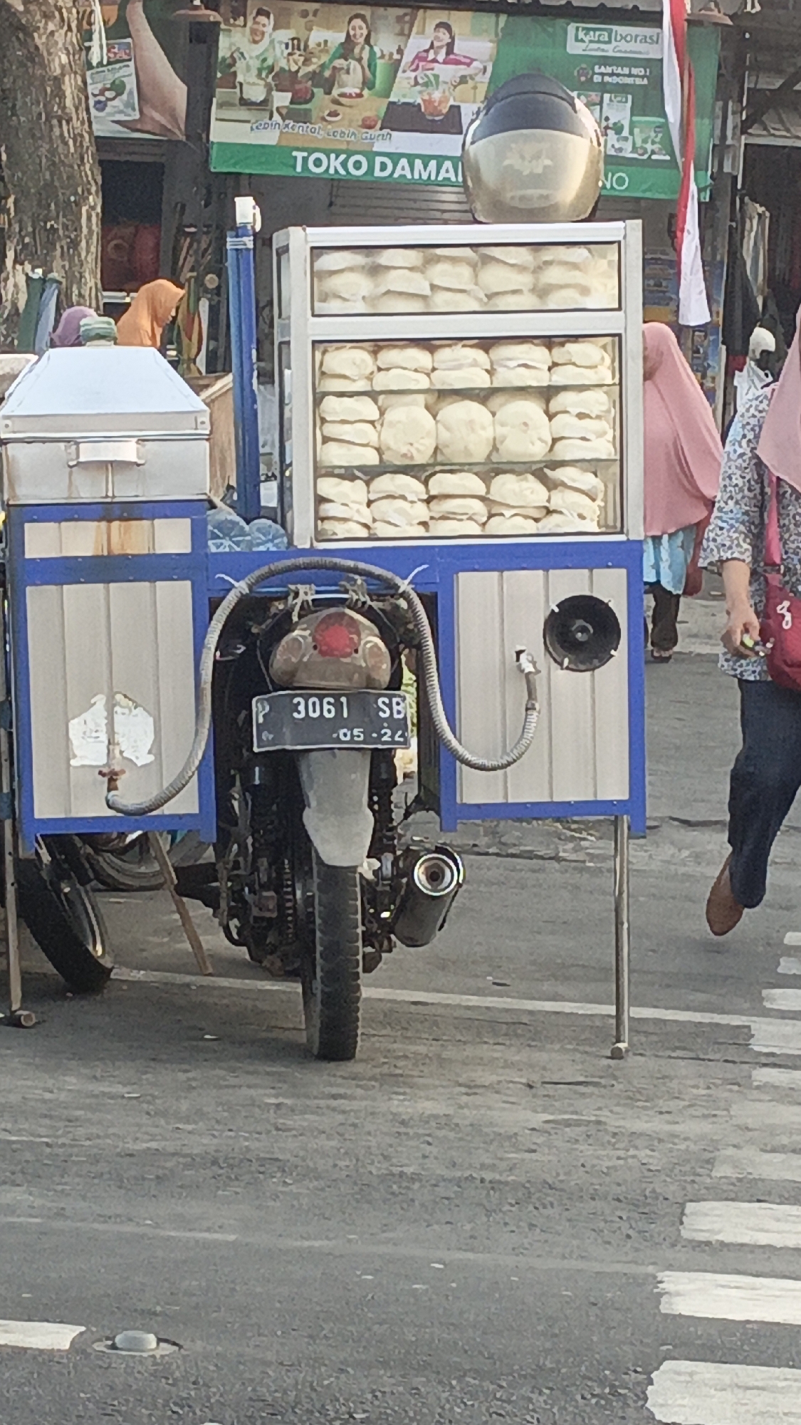 The image depicts a street scene in Srono, Indonesia, focused on a mobile food cart constructed from a motorcycle. The cart is filled with numerous round, white steamed buns, neatly arranged in a glass display case. The motorcycle's rear is visible, supporting the cart, with a license plate reading "P 3061 SB - 05-24". A person wearing a pink headscarf and a patterned blue and white top is seen walking away from the cart. Another person in an orange headscarf is partially visible behind the cart. A banner with green and white text, including "TOKO DAMAI", is displayed above the food cart. The environment appears to be an outdoor setting with a paved road and a pedestrian crossing.