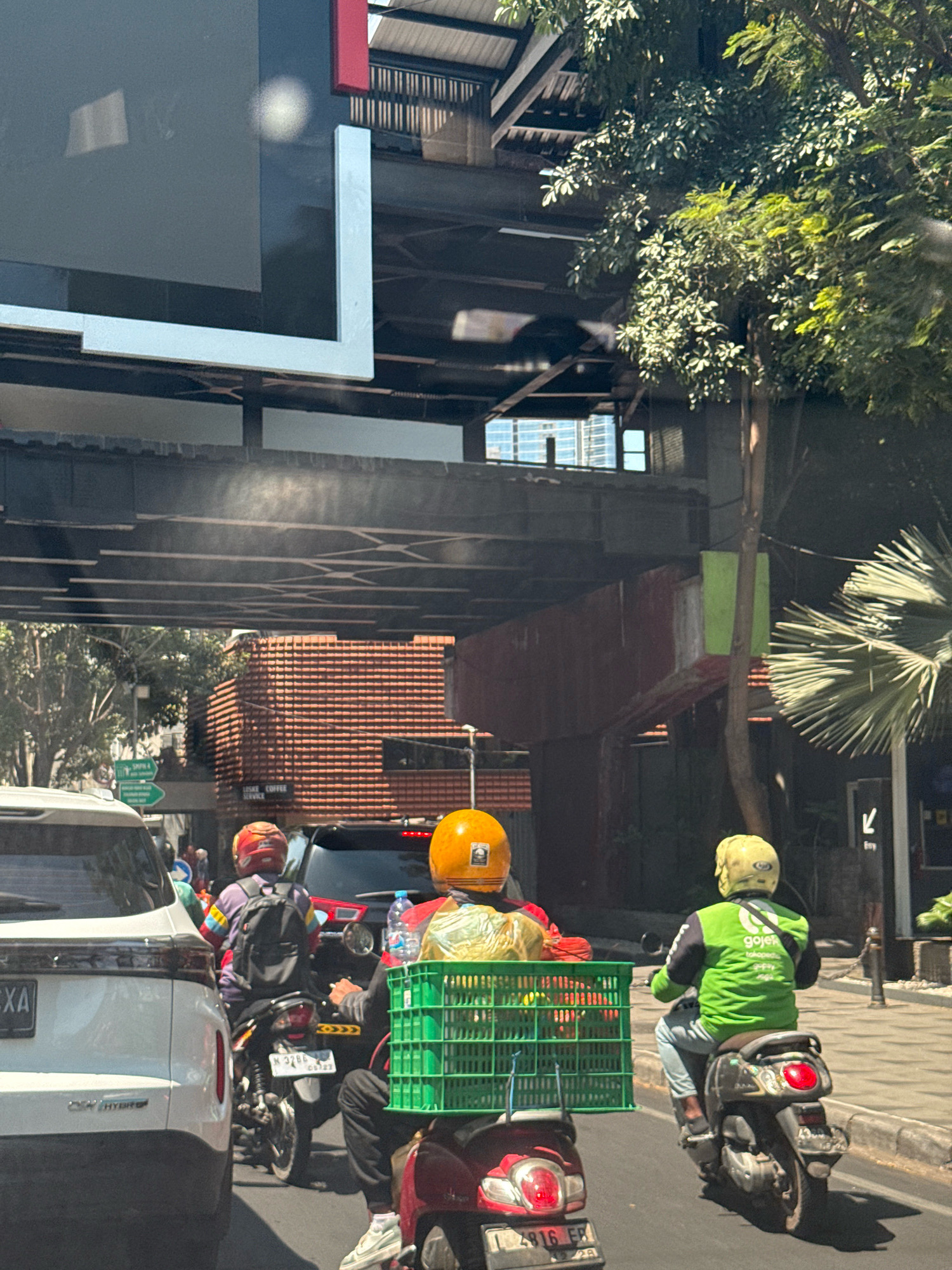 The image depicts a street scene in Surabaya, Indonesia, under an elevated structure, possibly a highway or overpass. In the foreground, a white SUV is partially visible on the left. Several motorcycles are in motion. One motorcycle, ridden by a person wearing a colorful shirt and a red helmet, has a backpack. Another motorcycle is ridden by a person in a green jacket with "Gojek" branding, indicating a ride-hailing service. This person is carrying a green plastic basket filled with items. A red motorcycle is also present. The environment shows buildings with tiled roofs and trees, suggesting an urban setting. The lighting indicates it is daytime, with strong sunlight casting shadows. Signs are visible in the background, including one with arrows and text that appears to be in Indonesian. The overall impression is a busy street with common modes of transportation and commercial activity.