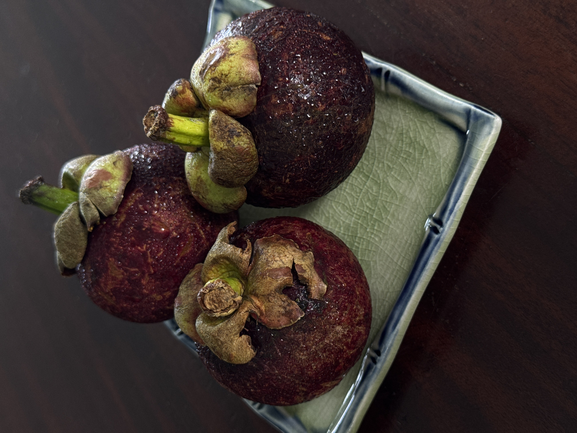 Three ripe mangosteens are arranged on a light green ceramic rectangular plate with a blue rim. The plate rests on a dark wooden surface. The mangosteens are a deep purple color with prominent green sepals at the top, and they are speckled with tiny water droplets. The background is dark and out of focus, providing a stark contrast to the brightly lit fruit. No people or specific locations within Goviyapana, Sri Lanka are depicted.