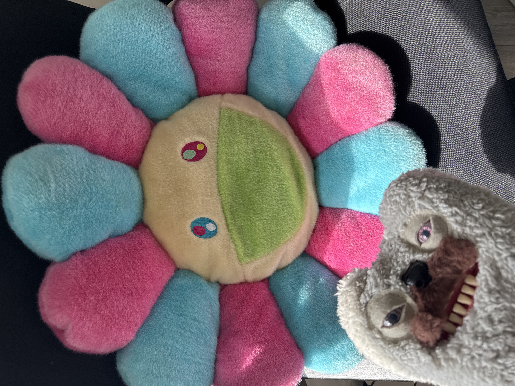The image shows two plush toys on a surface of dark and light gray material. On the left is a flower-shaped plush with alternating pink and light blue petals, and a split-colored face with one side beige and the other light green. The face has two sets of cartoon-like eyes. To the right is a light gray plush toy with a dark nose, and open mouth revealing yellow teeth. The background is blurred, but suggests an indoor setting. The location, Moscow, Russia is not discernible from the image.