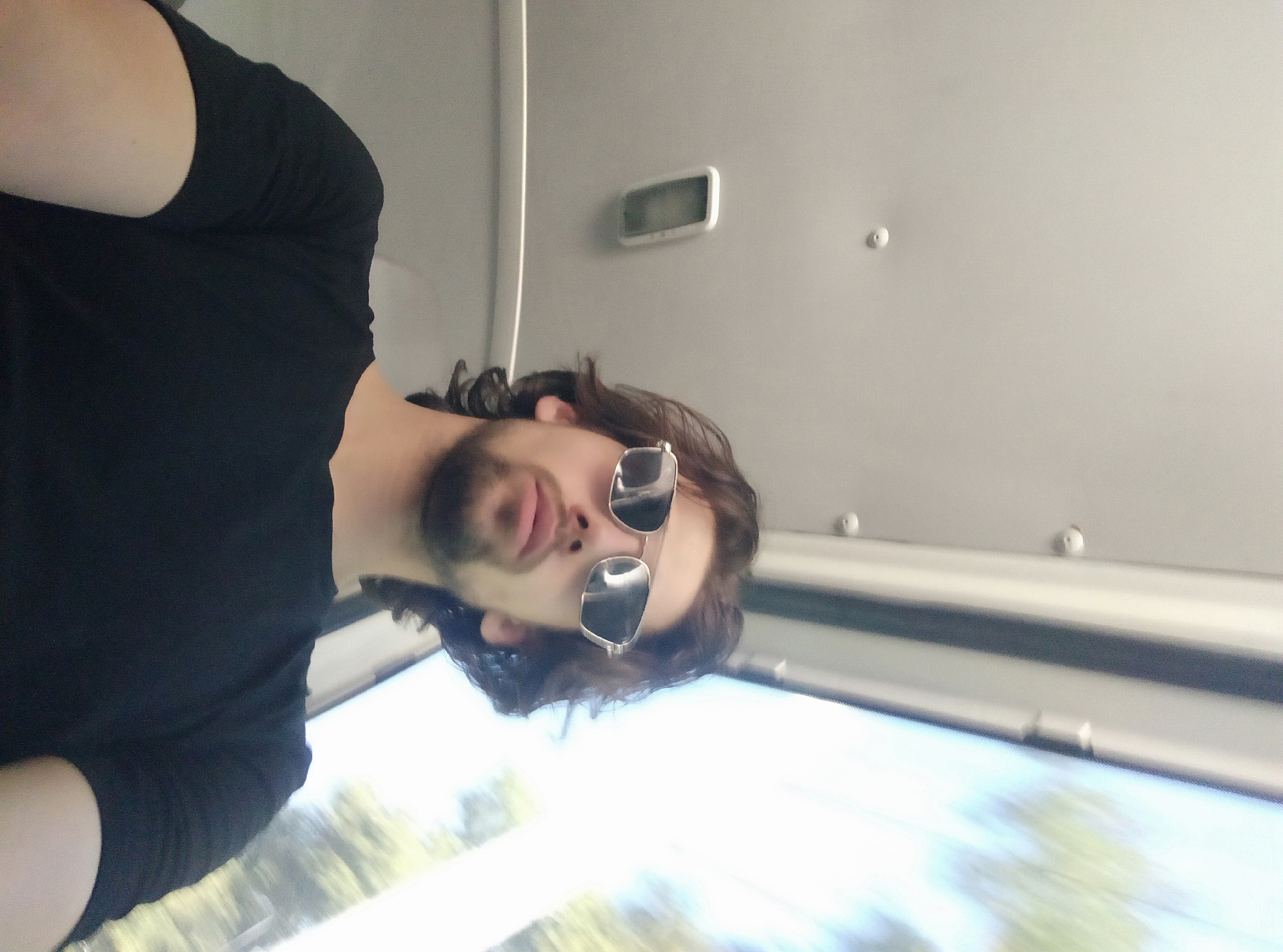 An underexposed, wide-angle selfie is taken from a low perspective, looking up at a young man with medium-length brown hair and a short beard. He is wearing a black t-shirt and silver-rimmed sunglasses with dark lenses. The background is blurry and suggests an indoor setting with a ceiling that has lights and what appears to be a window or opening showing blurred green foliage and a bright sky, indicative of daylight. The man is positioned slightly off-center to the left of the frame. The overall impression is of a casual portrait taken on the move.