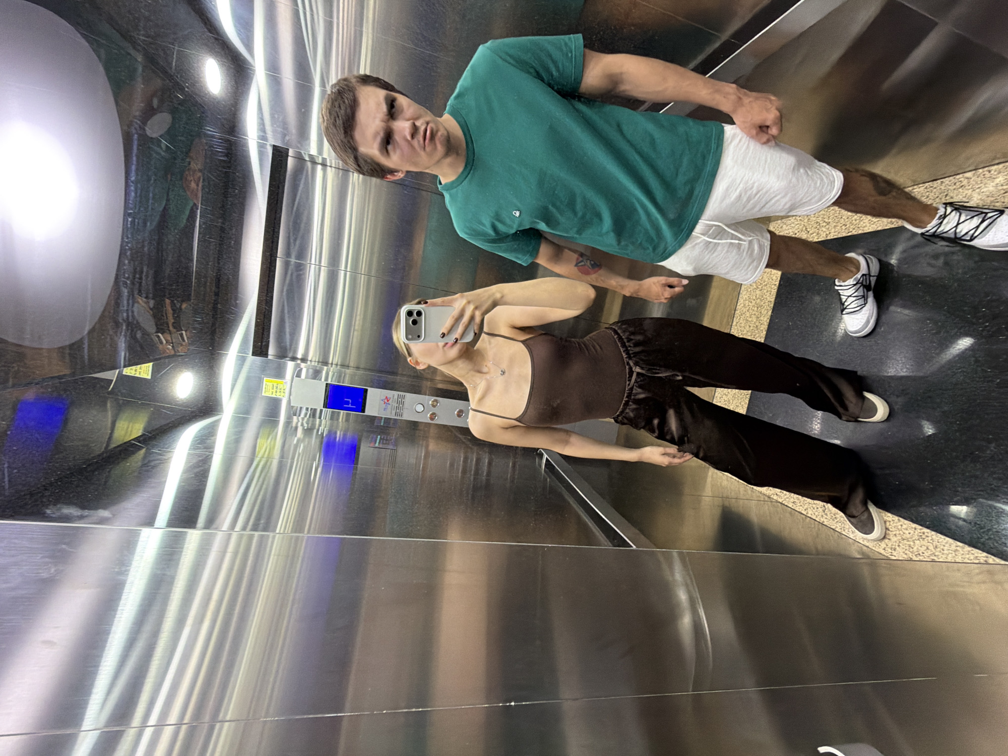 In an elevator with brushed metal walls, a young woman and a young man take a selfie. The woman, on the left, wears a dark brown tank top and loose, dark brown satin pants. She holds a smartphone in her left hand to capture the image, with her face tilted towards the device. A silver necklace with a small pendant is visible around her neck. Her right hand rests on her hip, and she wears white-soled sneakers with dark laces. The man, on the right, is in a green t-shirt and white shorts. He has dark hair and an intense, squared expression, looking directly at the camera. He wears white sneakers with black laces. The elevator floor has a speckled pattern transitioning to dark, reflective black. The lighting is bright, reflecting off the metallic surfaces of the elevator. The context of Đà Nẵng, Vietnam is not discernible from the visual elements of the image.