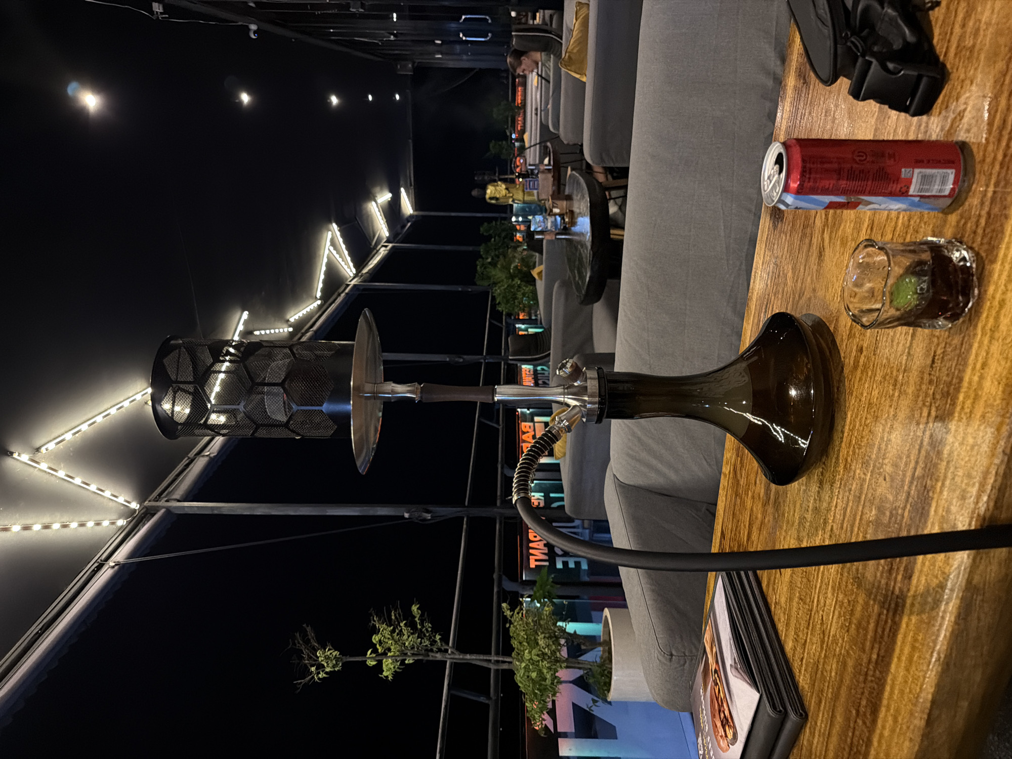 The image depicts an outdoor setting in North Kuta, Indonesia, featuring a wooden table with a hookah, a partially visible can of beer, and a glass with a dark liquid and a green garnish. A black backpack rests nearby. The setting includes gray upholstered seating. In the background, a person is visible seated on similar upholstery. Further back, there are potted plants and illuminated elements, potentially signage or lighting structures. The scene is likely at night due to the darkness and the presence of artificial lights.