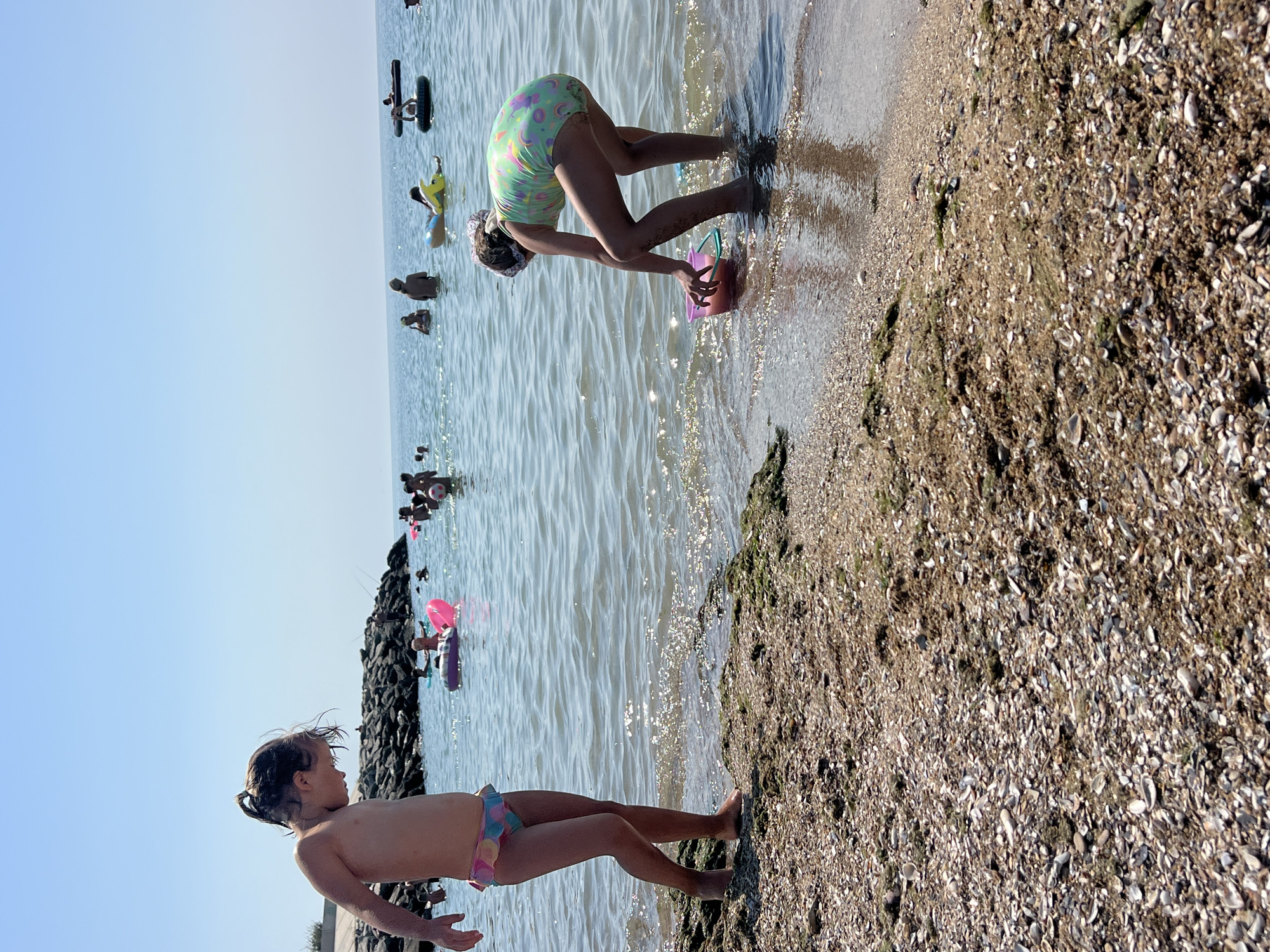 The image shows a beach scene on a sunny day. In the foreground, a young girl with wet hair is standing near the water's edge, looking to her right. She is wearing a colorful swimsuit. Behind her, a second young girl in a patterned swimsuit is bent over at the edge of the water, holding a pink bucket and playing. Several other people are visible in the water in the background, some on inflatables. The water is calm and reflects the bright sky. The beach itself is covered in sand and small shells. The scene suggests a leisurely day spent at the seaside. There is no visible text in the image. The location context of Fontanka, Ukraine, indicates this is likely a Black Sea beach.