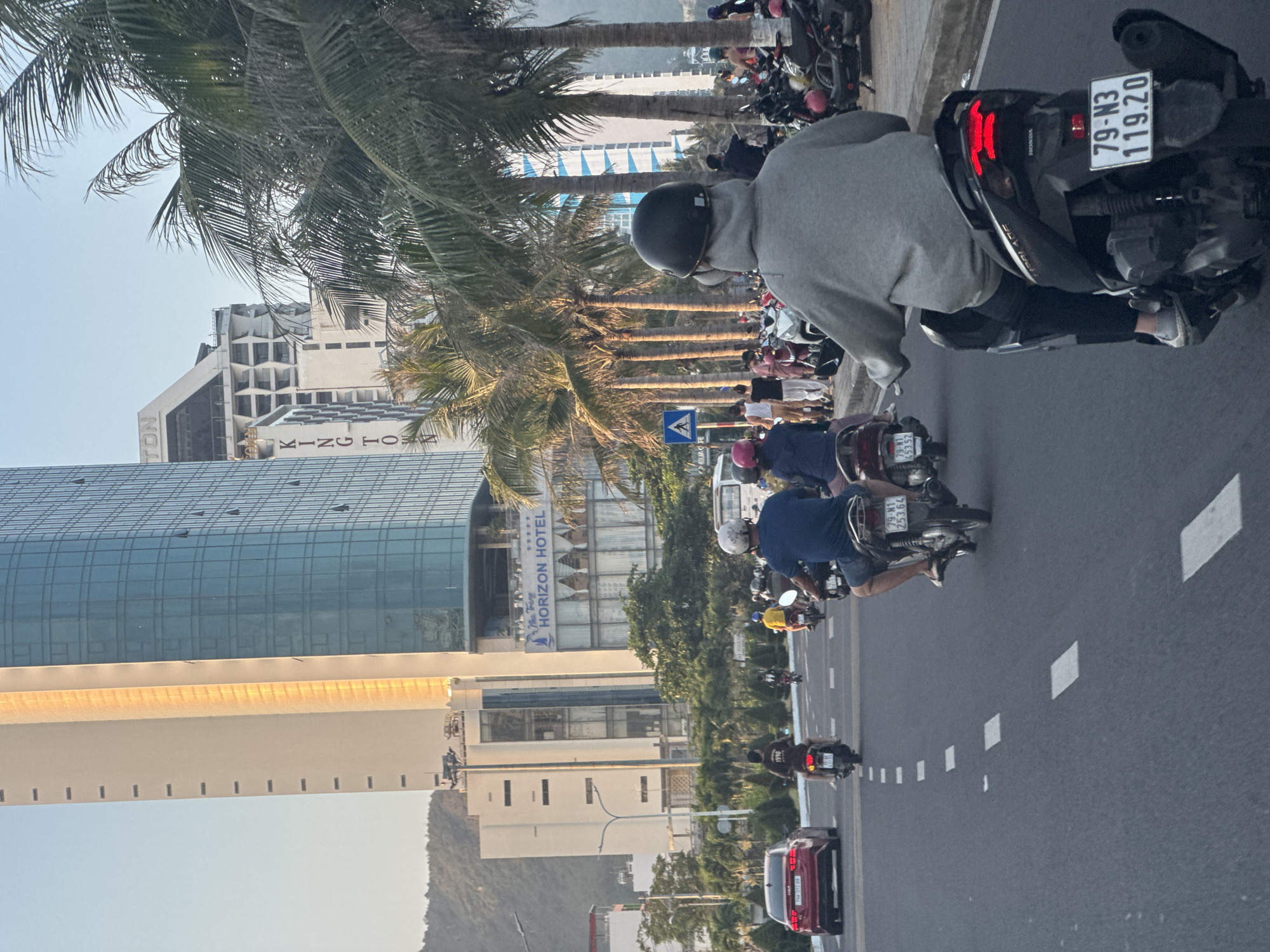 The image depicts an urban street scene in Bac Nha Trang Ward, Nha Trang, Vietnam, during what appears to be late afternoon or early morning with clear skies and bright natural light. The primary focus is a multi-lane asphalt road populated by numerous motorbikes and one red sedan, with an active sidewalk and prominent buildings lining the right side.
In the foreground, the back of a person on a black Honda motorbike with a visible license plate "79-N3 119.20" is seen. This rider wears a black helmet, a grey hooded sweatshirt, and dark pants. The motorbike's red tail lights are illuminated. To the rider's left, a person on another motorbike with license plate "79-M1 453.52" is partially visible. Further down the road, additional motorbikes are in motion, including one with license plate "79-M1 253.64". A red sedan, possibly a Mazda 3, with license plate "79A-372.09", is positioned in the left lane. All motorbike riders visible in detail are wearing helmets. The road features white dashed lane markings.
To the right of the road, a wide paved sidewalk is lined with numerous tall palm trees, casting long shadows to the left. Many motorbikes are parked perpendicularly along this sidewalk. Pedestrians are observed walking on the sidewalk amongst the parked vehicles and palm trees; a blue pedestrian crossing sign is visible on a pole near the sidewalk.
Beyond the palm trees and sidewalk, several modern high-rise buildings dominate the background. Prominently visible is the "Nha Trang HORIZON HOTEL" sign, indicating a five-star rating, situated on a large building with glass and white architectural elements. Another distinctive building partially obscured by palm trees bears the vertical red lettering "KINGTOWN" and features an angular roof design and patterned facade. Another tall building exhibits a vertical strip of intense golden-orange reflection, likely from direct sunlight. In the far background, on the left side of the image, green mountains are visible under the clear sky.