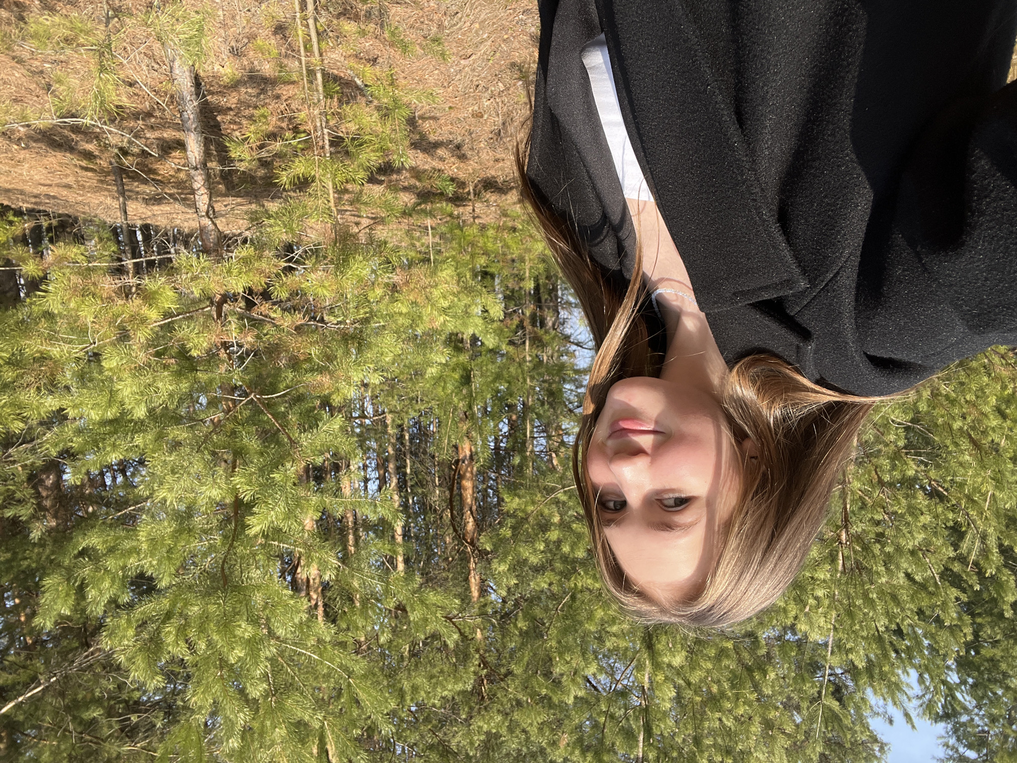 A woman is in a coniferous forest located in Chaikovsky, Russia. She is wearing a black coat and a white shirt. The forest consists primarily of pine trees. The ground is covered with pine needles. The sky is visible through the trees.