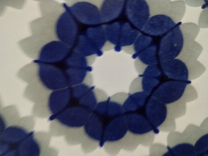 The image shows a pattern of stylized floral elements arranged in circles. The "petals" of each flower are blue, with short lines extending outward from the centers of the intersections. The backgrounds around the flowers are white, with a soft, muted gray outlining the floral shapes, creating a blurred, shadow-like effect. The overall impression is a repeated, stylized floral design on a surface.