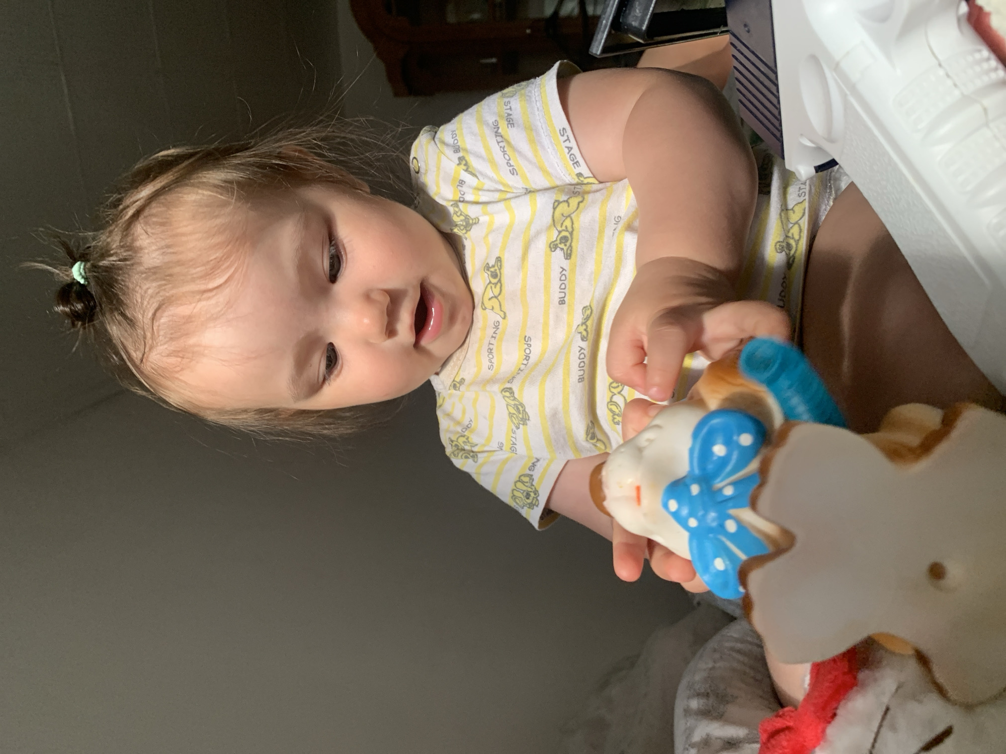In a dimly lit room, likely a bedroom given the soft furnishings and clothing, a young child, approximately 1-2 years old, is seated and actively engaged with toys. The child has light brown hair pulled into a small bun on top of their head with a green hair tie and is wearing a yellow and white striped t-shirt adorned with a repeating pattern of cartoon dogs. The child's gaze is directed downwards, focused on a stuffed toy, possibly a bunny, held in their hands. The toy has a blue bow with white polka dots and appears to be made of soft material. To the child's right, a white toy gun is visible, partially obscuring the child's lower body. The background is dark and out of focus, with a hint of a wooden cabinet or furniture on the right side of the frame. The overall impression is one of a quiet moment of play.
