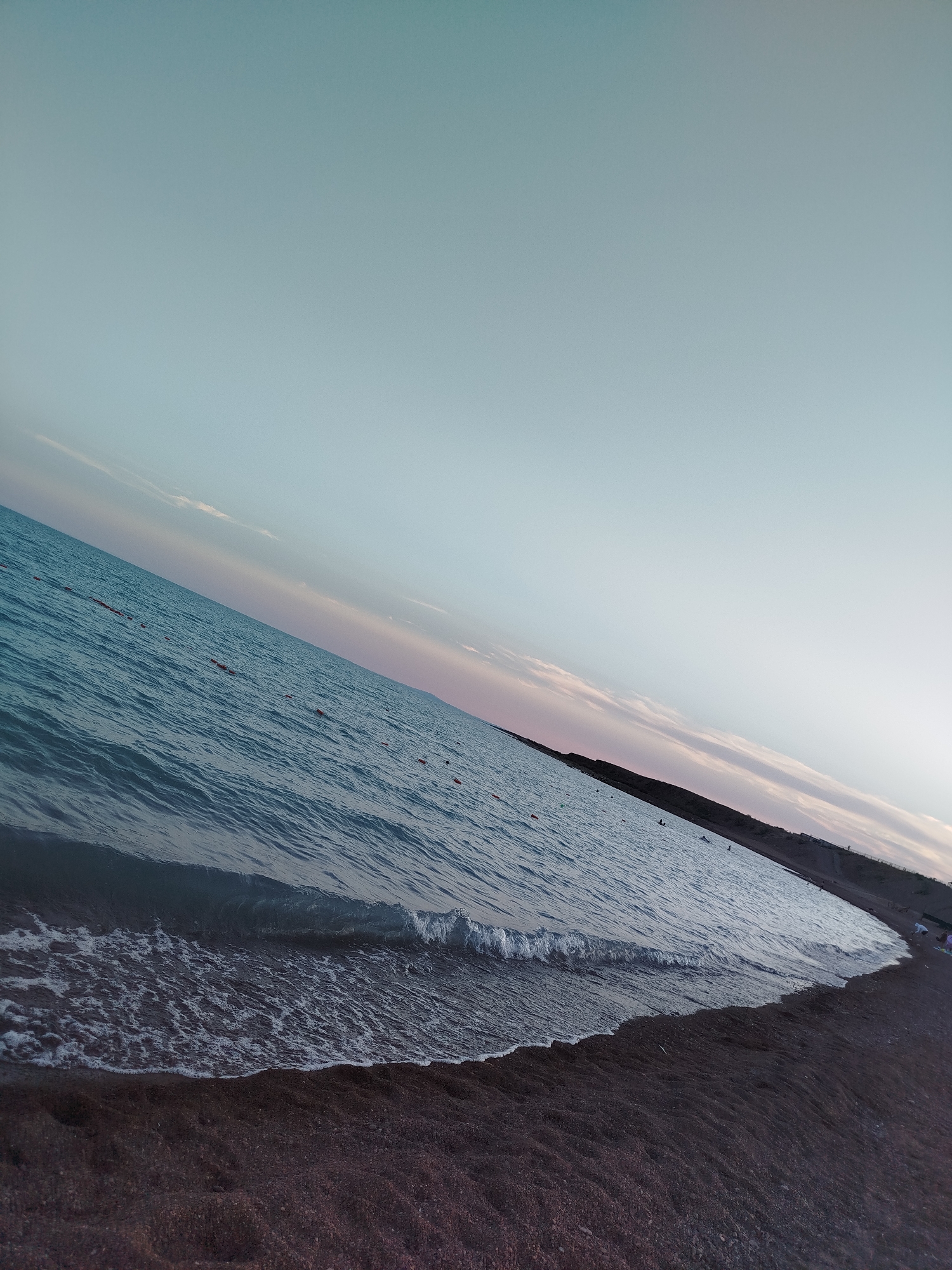 The image depicts a serene beach scene at what appears to be either sunrise or sunset. The perspective is angled, with the shoreline running diagonally from the bottom right to the upper left of the frame. The ocean occupies the majority of the image, with calm, rippling water in shades of deep blue and teal. A gentle wave is breaking on the shore in the foreground, creating a foamy white edge. The beach itself is composed of dark, fine-grained sand. In the distance, a dark, sloped landmass is visible along the horizon, possibly a coastline or hills. The sky is a gradient of soft blues and pale pinks, with faint, wispy clouds near the horizon. A few small, orange buoys are scattered in the water. A handful of distant figures, too small to discern details, are present on the beach and in the water, suggesting a lightly populated area. The overall atmosphere is tranquil and expansive.