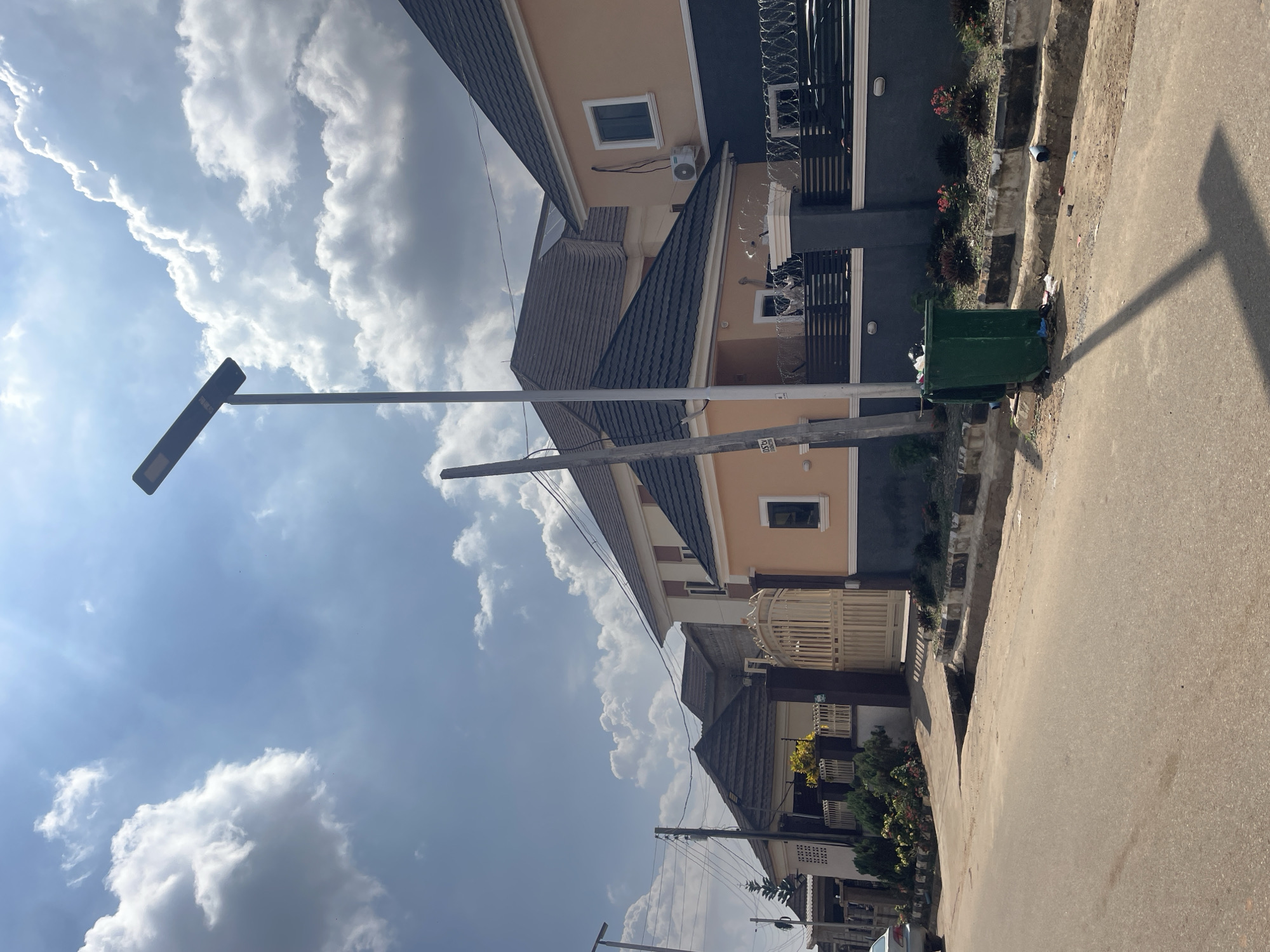 A street scene in Abuja, Nigeria, features a row of modern houses with pitched roofs and light-colored facades. A tall solar-powered street lamp stands prominently on the right side of the image, casting a shadow across the asphalt road. A green municipal waste bin is positioned on the sidewalk near the street lamp, with visible debris around it. A beige metal gate, adorned with decorative patterns, is visible further down the street, leading into one of the properties. The sky is partly cloudy with the sun creating bright patches. A silver car is parked on the left side of the road.