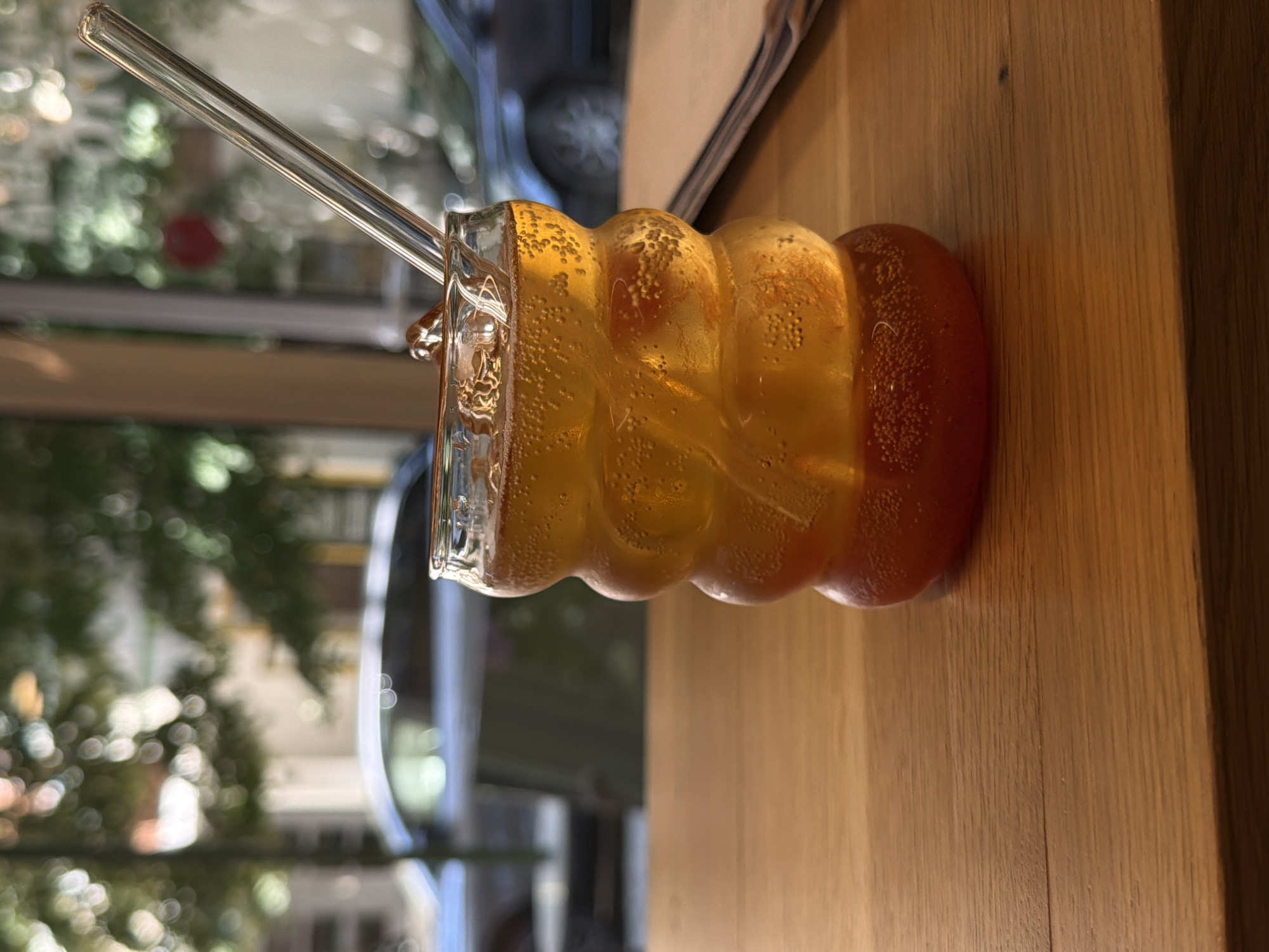 A close-up, eye-level, indoor shot focuses on a bubbly amber-colored beverage in a uniquely shaped, ribbed glass with a glass straw. The drink is filled with ice cubes and appears to be carbonated. The glass sits on a wooden table. In the blurred background, hints of a city street are visible through a window, with greenery and the shapes of cars, suggesting an urban setting like Sofia, Bulgaria. The overall lighting is natural, possibly from the window. There are no people or specific actions depicted, only the still life of the drink and its surroundings.