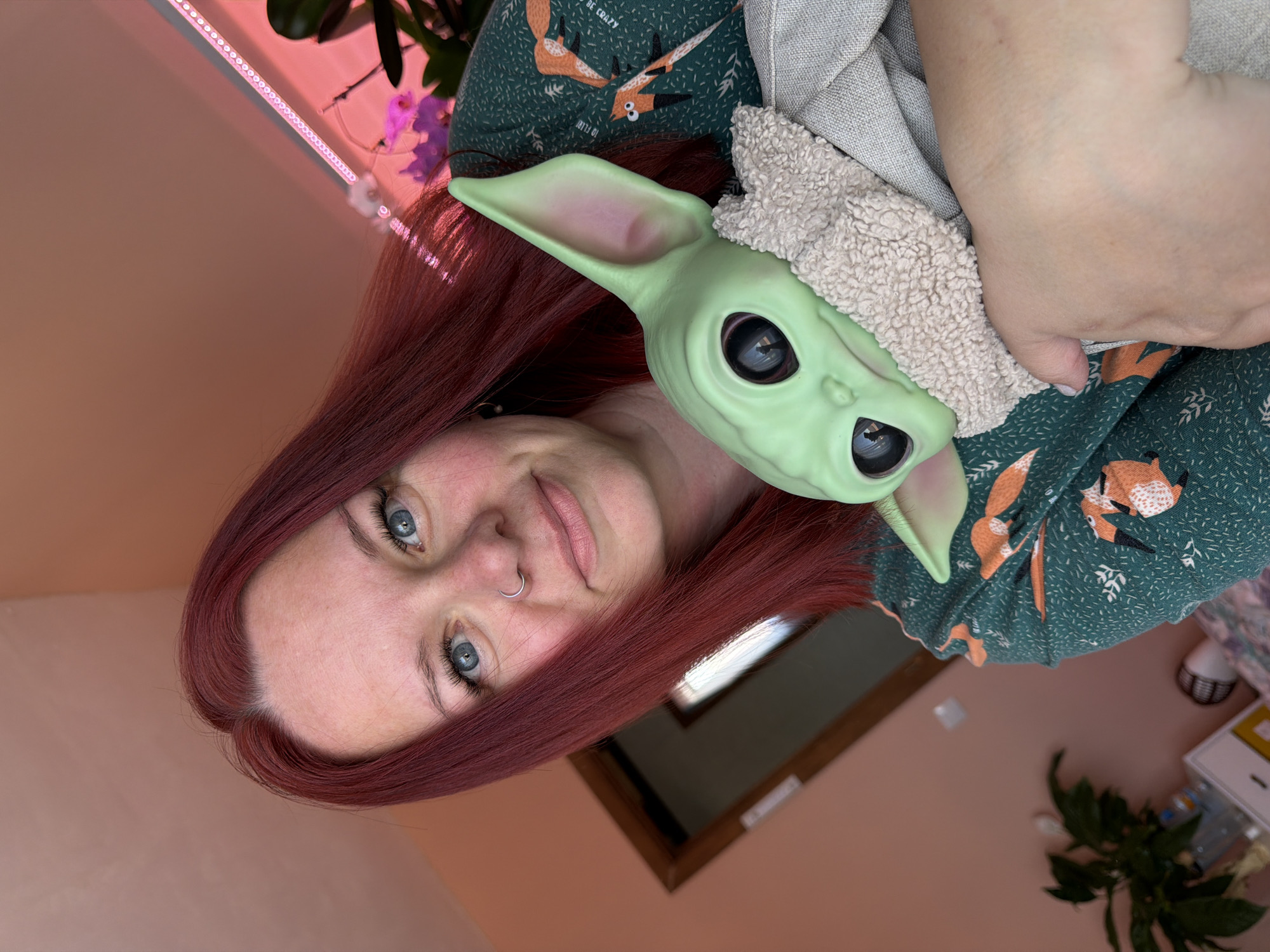 A medium shot shows a light-skinned woman with red hair holding a Grogu (Baby Yoda) doll wearing a light brown hat. She has blue eyes, a nose ring, and is looking at the camera with a slight smile. She is wearing a green dress with a fox pattern. The background includes a pink wall with an LED light strip, a plant with orchids, a brown-framed window, another plant, and a white desk with objects on it. The location is implied to be in Chaikovsky, Russia, but this is not verifiable from the image.